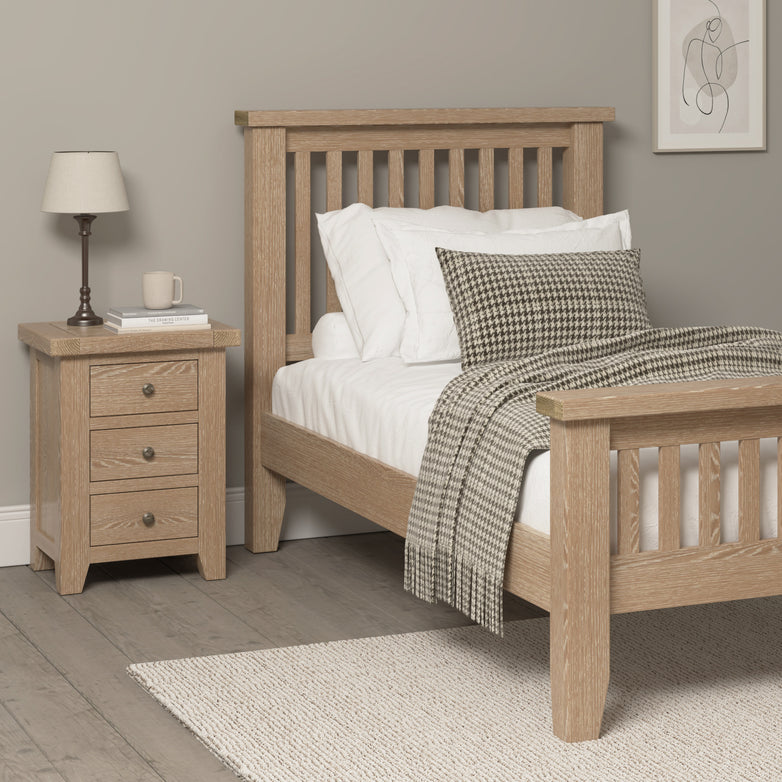 Wooden bed and the Burford 3 Drawers Bedside Table in Natural Oak with lamp in a bedroom; bed with white linens, patterned throw, and cushion; modern decor featuring ample storage.