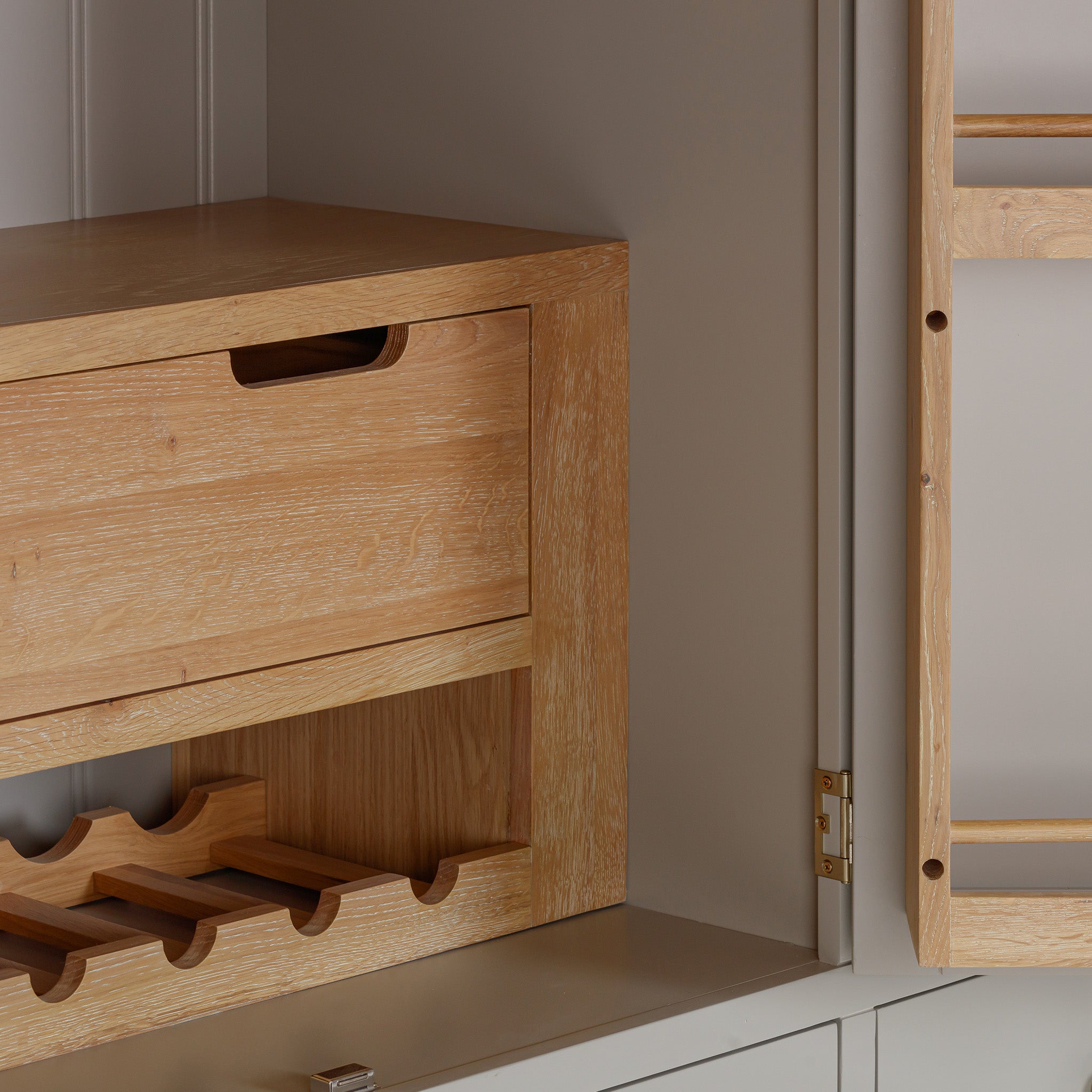 The Burford Triple Larder Unit in Pebble Grey features a natural oak finish, functioning as both a wine rack and cabinet with a small drawer. It is elegantly positioned against a light-colored wall, serving as an ideal kitchen storage solution.