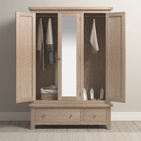 The Burford Triple Wardrobe with 2 Drawers in Natural Oak is the perfect storage solution, featuring open doors that reveal hanging clothes, a central mirror, and a basket on the right shelf. Made from natural oak, this piece combines elegance with functionality.