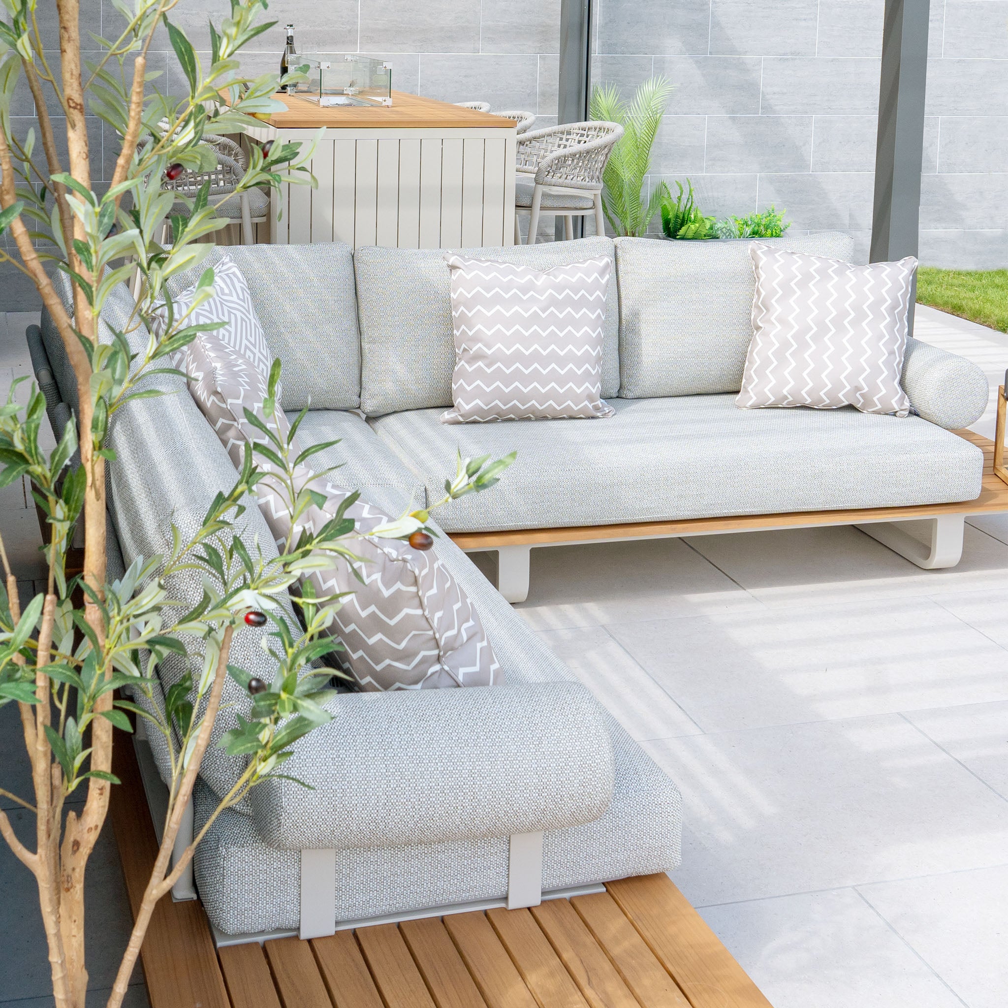 Modern outdoor patio featuring a Bay Corner Group Set with 4 Waist Pillows and a Teak Table in Latte, showcasing a gray sectional sofa adorned with chevron pillows and wooden accents. The sectional boasts a sleek powder-coated aluminum frame and durable Olefin material for lasting comfort and style, with the dining table visible in the background.