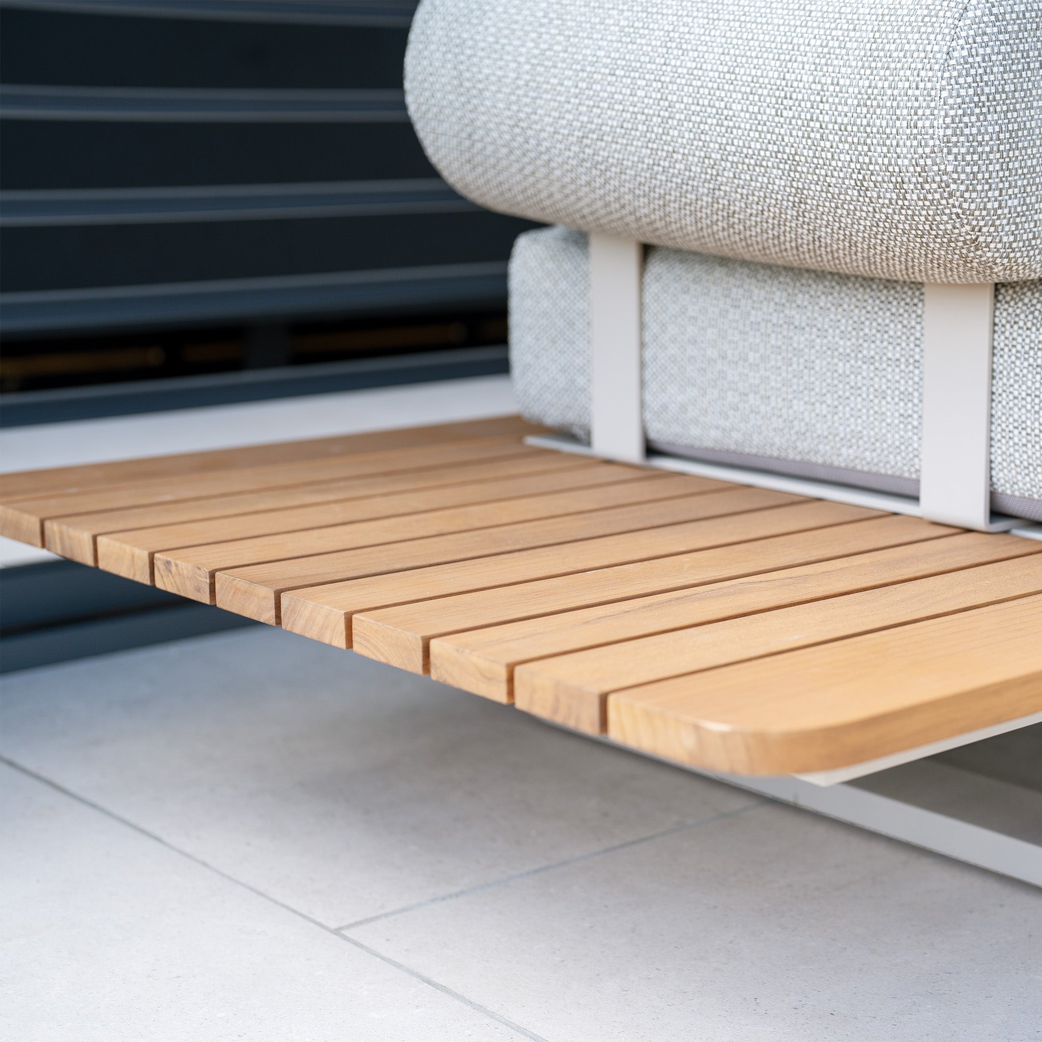 Close-up of the Bay Corner Group Set with 4 Waist Pillows Teak Table in Latte, showcasing its modern wooden slatted bench with gray cushions, set on a light tile floor, and featuring a durable powder-coated aluminum frame.