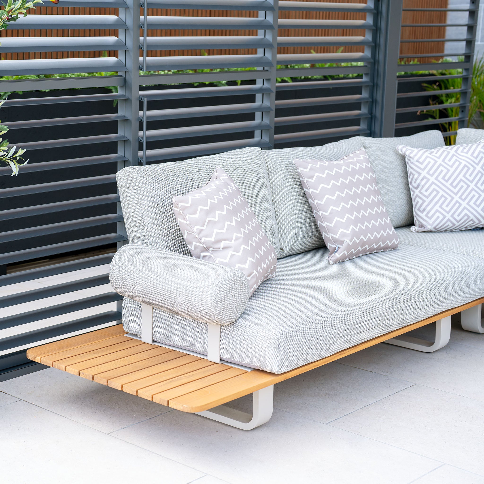 The contemporary Bay Corner Group Set with 4 Waist Pillows and a Teak Table in Latte, adorned with light grey cushions and patterned pillows, is showcased on a patio featuring a privacy screen with grey horizontal slats and boasts a durable powder-coated aluminium frame.