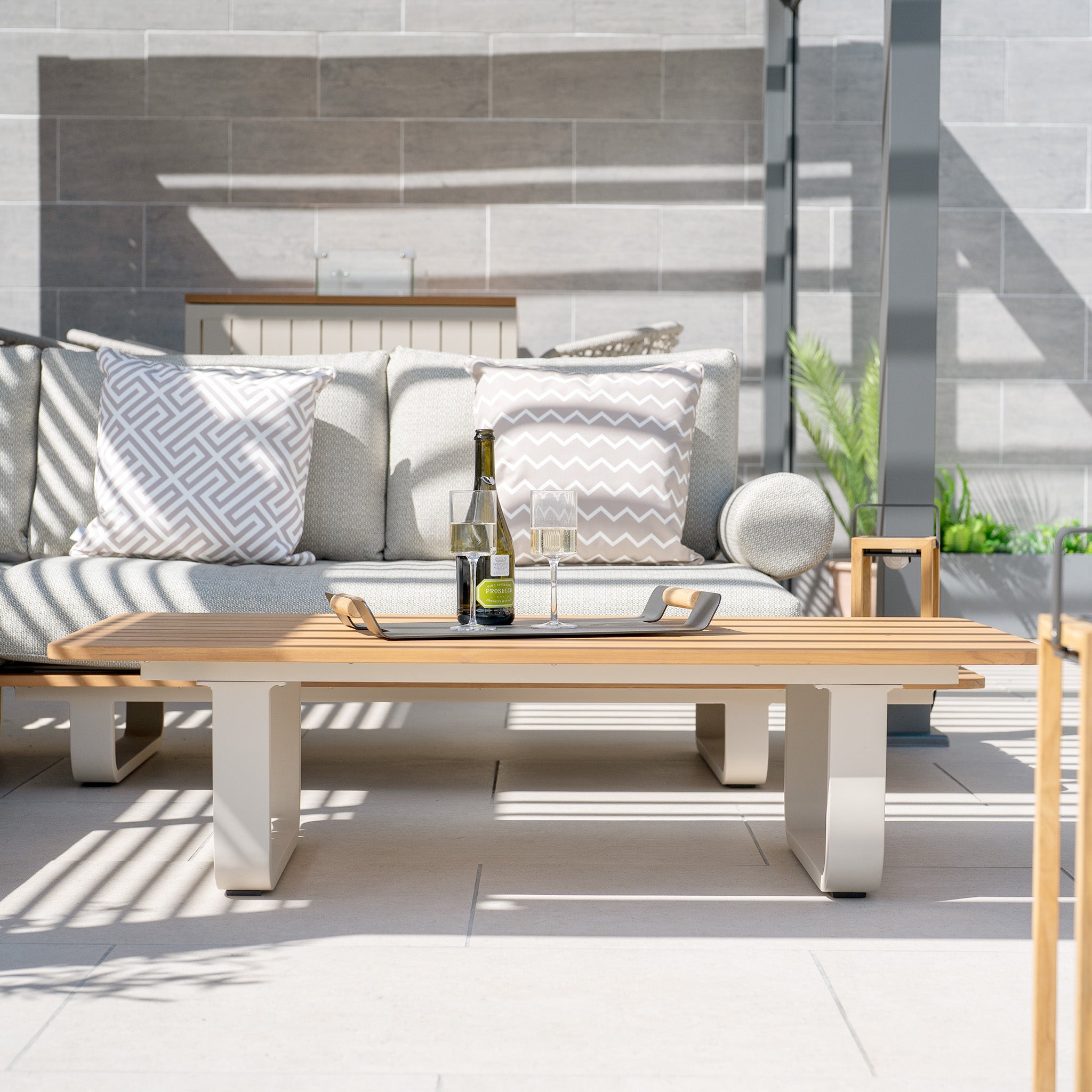 Modern outdoor lounge area with a teak table, wine bottle, and two glasses. Sofa from the Bay Corner Group Set with 4 Waist Pillows in the background, featuring a sturdy powder-coated aluminium frame for durability.