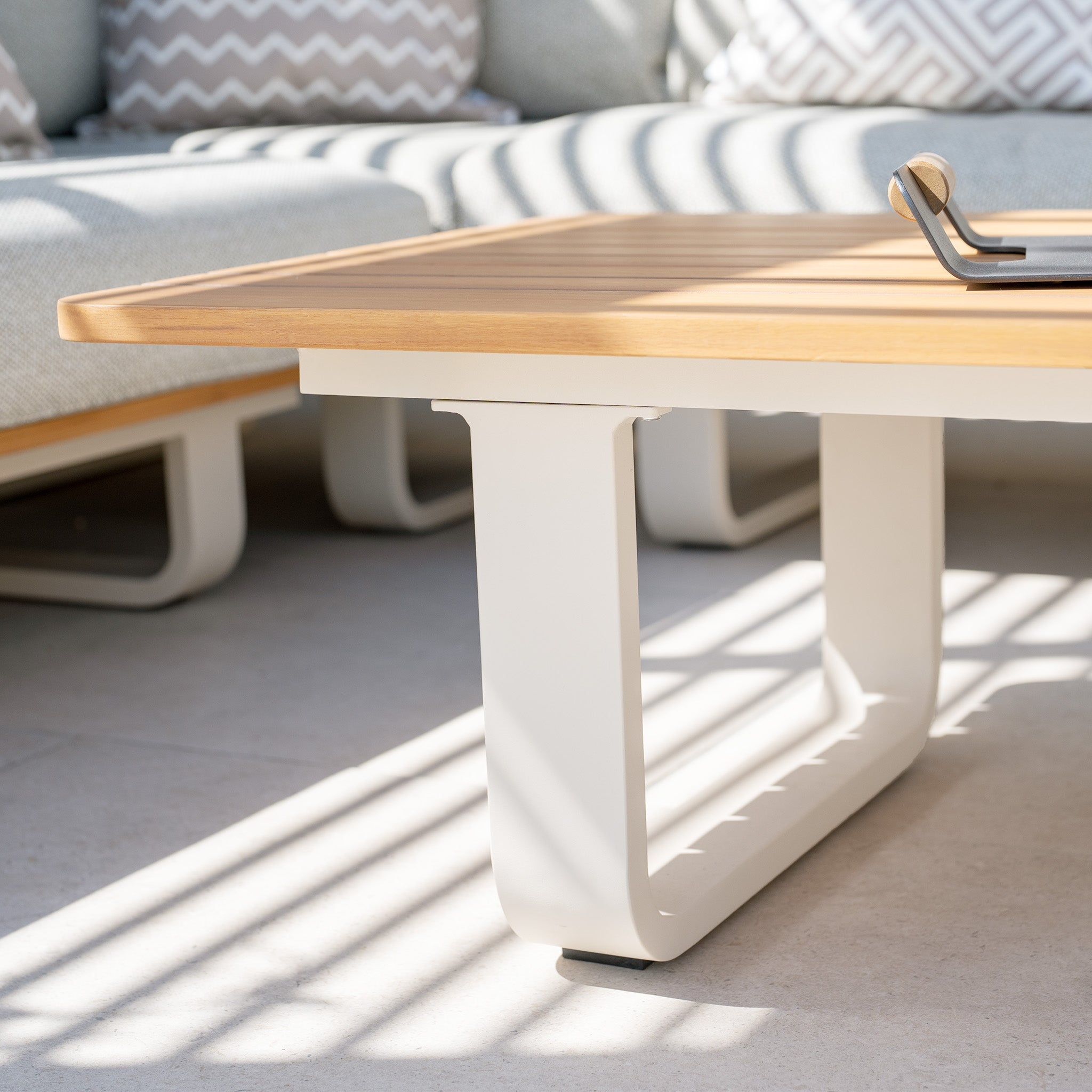 A modern light wood coffee table featuring white U-shaped legs sits on a sunlit patio, surrounded by patterned cushions. The Bay Corner Group Set with 4 Waist Pillows and a Teak Table in Latte is crafted from Olefin material, adding durability and elegance to your outdoor setting.