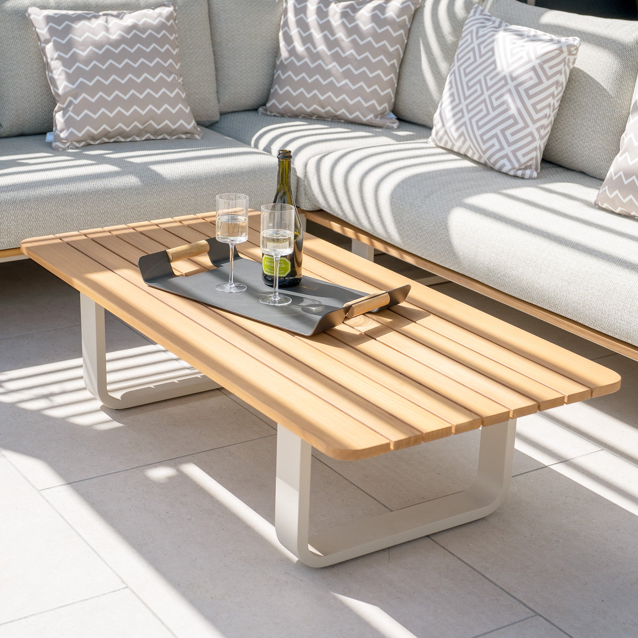 Bay Corner Group Set with four waist pillows in Latte, featuring a teak table holding drinks on a tray, surrounded by a pale gray sectional couch made from Olefin material and accented with patterned cushions.