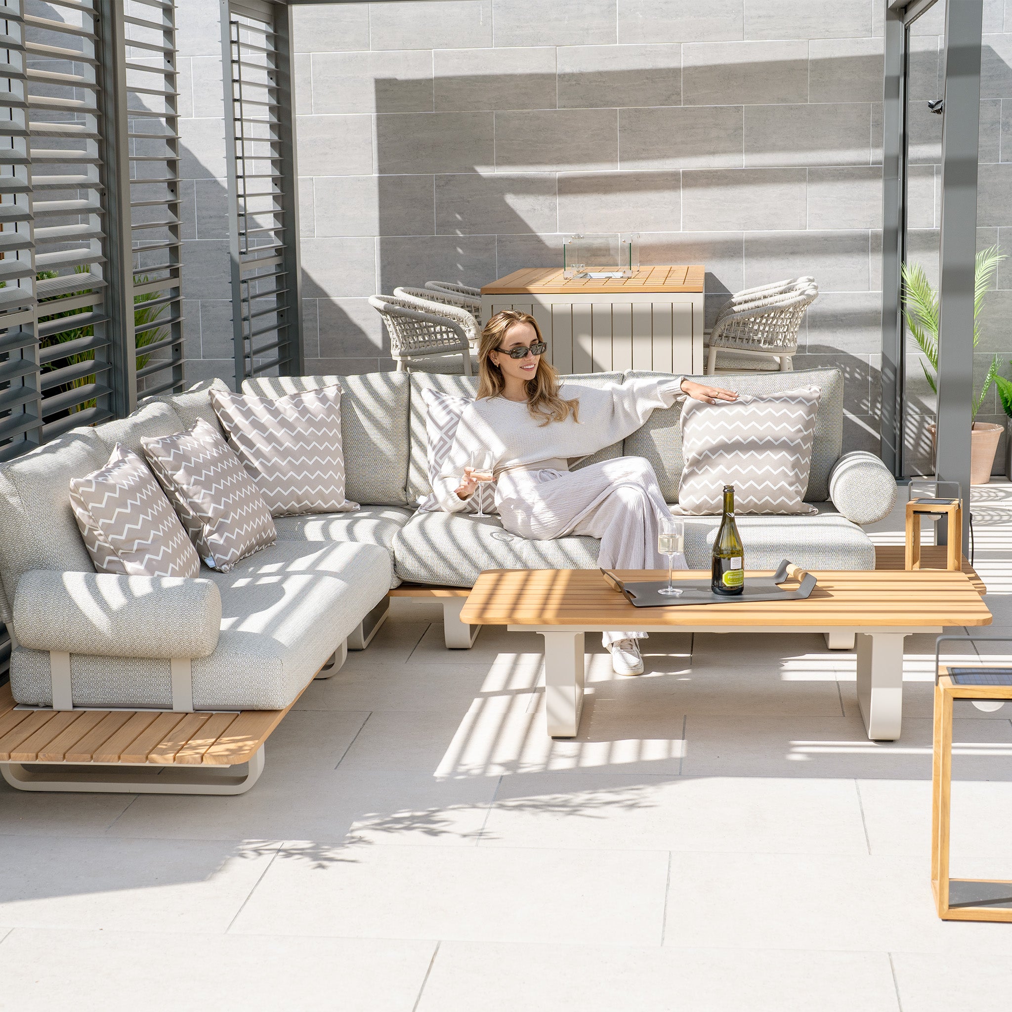 A woman in white sits on a light grey outdoor couch with patterned pillows under a pergola, featuring the Bay Corner Group Set with 4 Waist Pillows and a Teak Table in Latte. A bottle and glass rest on the table, showcasing the powder-coated aluminum frame—ideal for any outdoor setting.