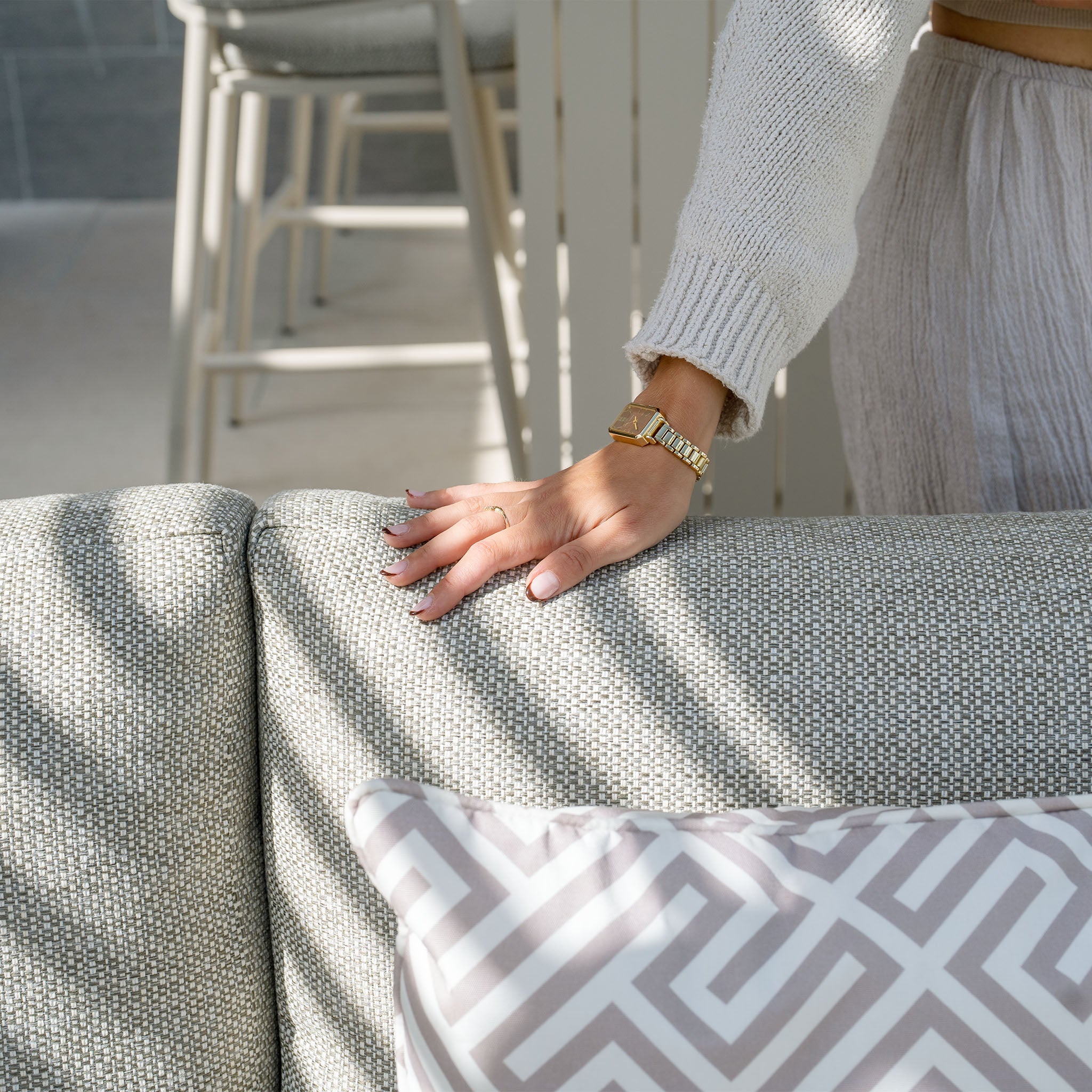 A person wearing a sweater rests their hand on the back of a gray sofa with a decorative white and gray cushion, part of the stylish Bay Corner Group Set with 4 Waist Pillows Teak Table in Latte collection.