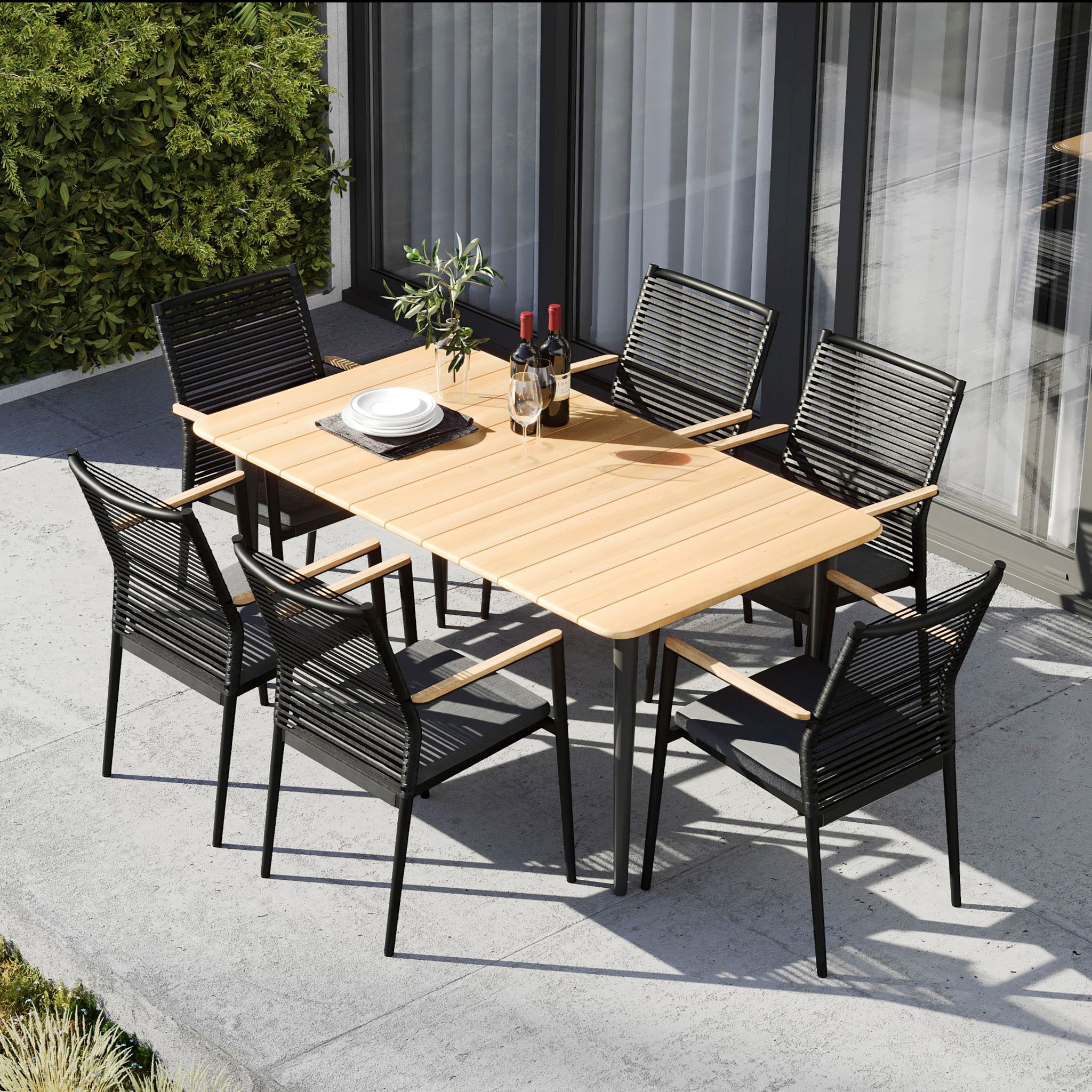 Introducing the Portland 6 Seat Rectangular Dining Set with Teak Table in Charcoal, featuring a teak table and six black chairs with weatherproof aluminum legs, complemented by a potted plant, all elegantly arranged on a concrete patio near lush greenery.