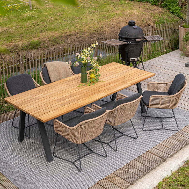 Outdoor dining setup with the Belmond 6 Seat Dining Set featuring a natural teak table, weatherproof wood, and six chairs, accompanied by a nearby grill and lush greenery in the background.