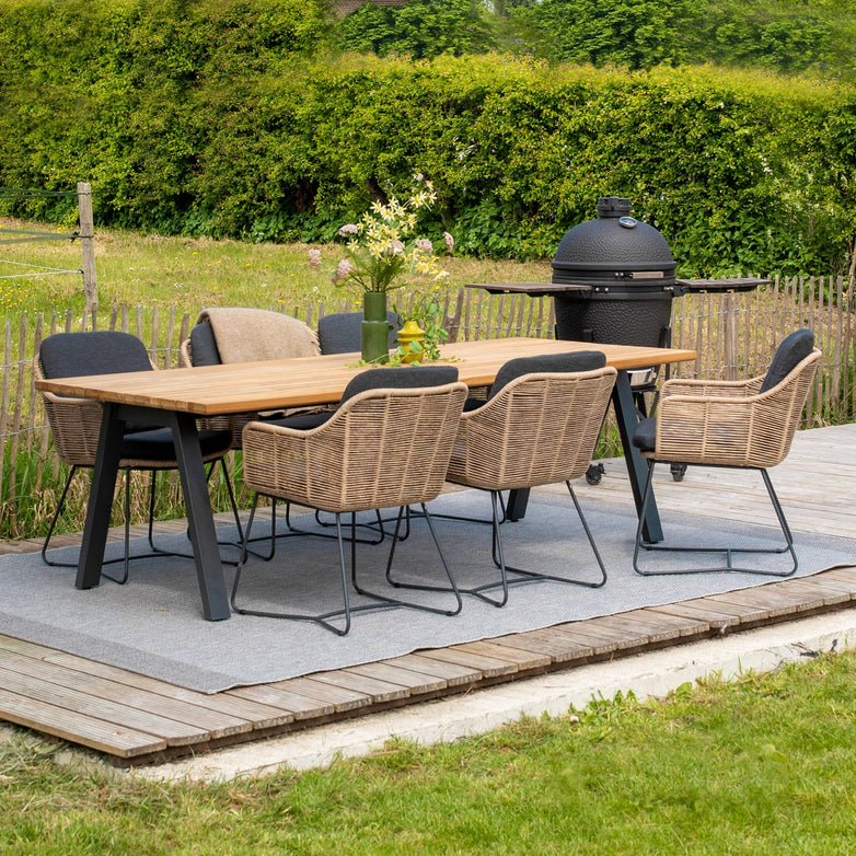 Outdoor dining area featuring the Belmond 6 Seat Dining Set with a natural teak table and six hand-woven natural poly rattan chairs, along with a black grill on a wooden deck surrounded by greenery. The weatherproof furniture ensures durability and timeless style through all seasons.