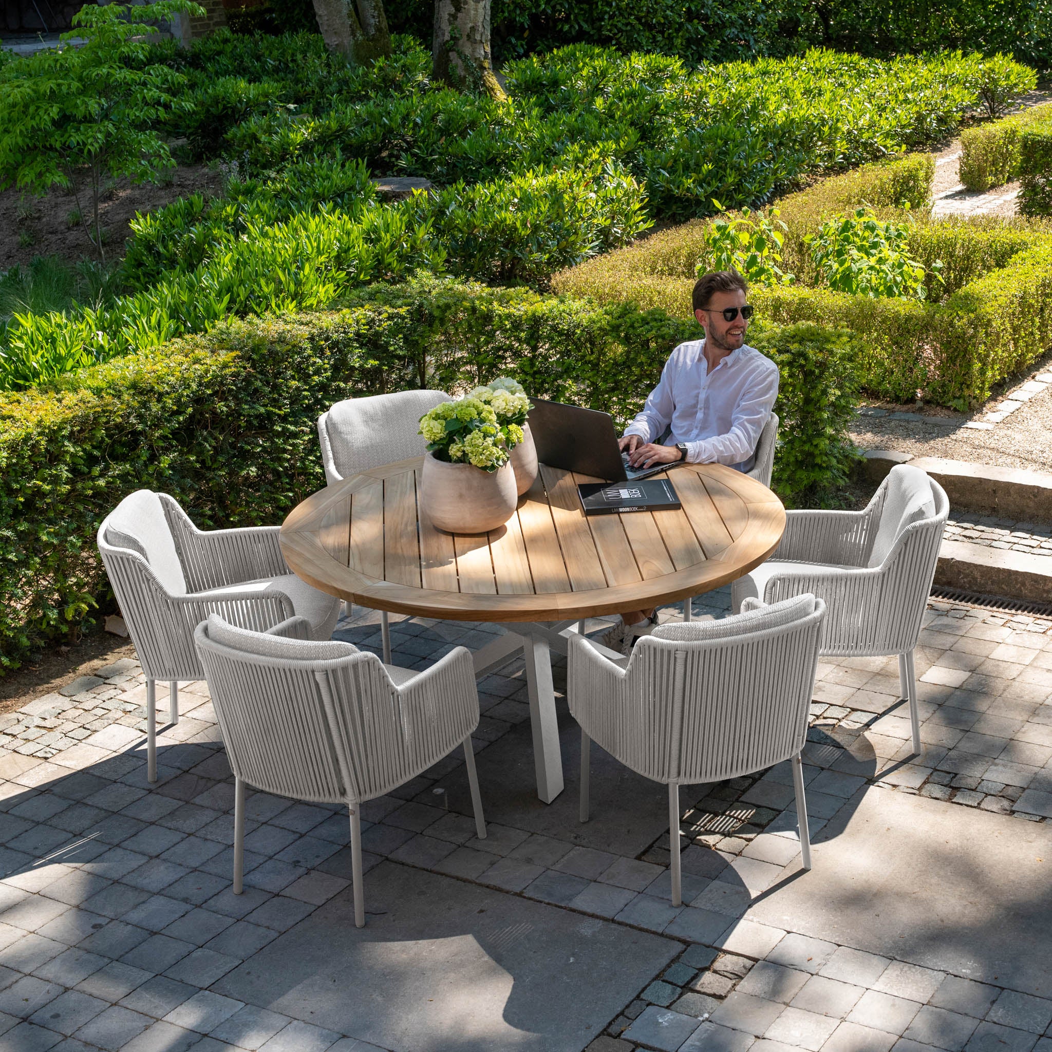 A man with a laptop sits at the Bernini 6 Seat Round Dining with Teak Table in Light Grey, accompanied by white chairs in a sunlit garden.