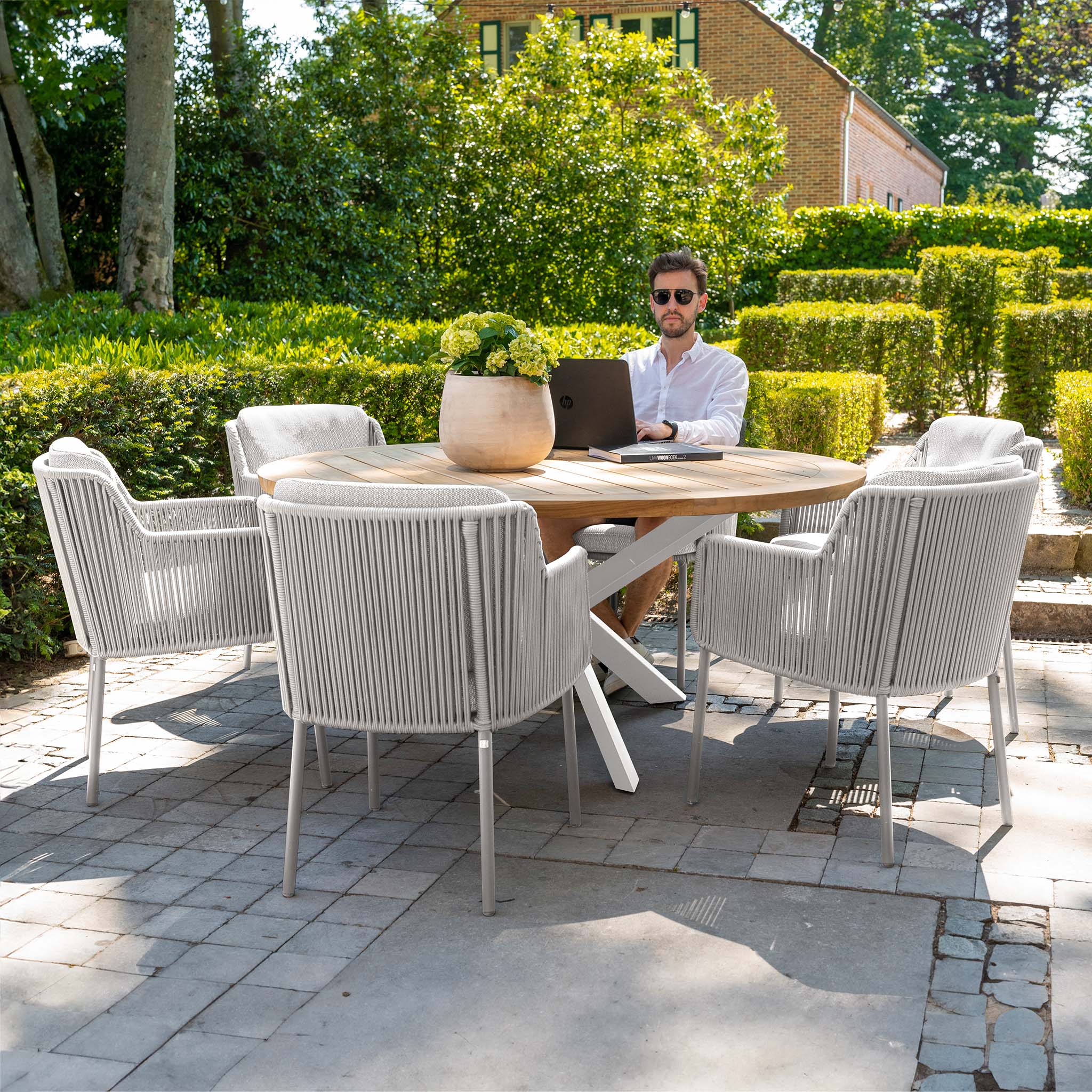 A man works on a laptop at a Bernini 6 Seat Round Dining with Teak Table in Light Grey, accompanied by six white chairs from the 4 Seasons Rope collection, surrounded by greenery and a brick building in the background. This elegant setup comes with a 5-year guarantee for peace of mind.