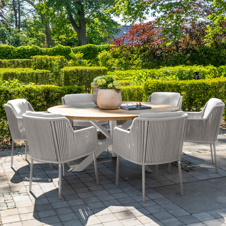 The Bernini 6 Seat Round Dining with Teak Table in Light Grey comes with six white chairs and a potted plant in the center, set on a stone patio surrounded by greenery. Enjoy peace of mind with our 5-year guarantee.