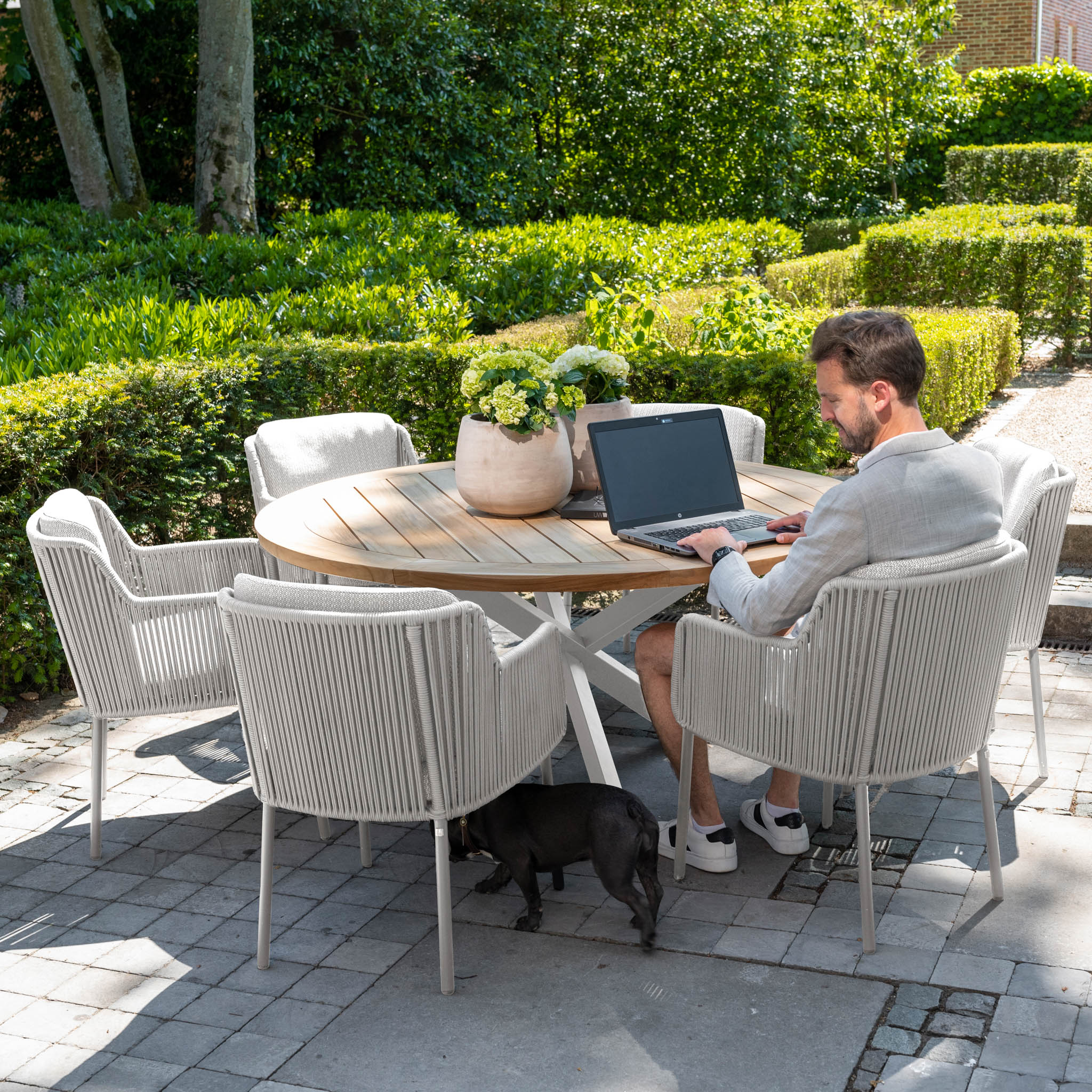 A man works on a laptop at a round outdoor table from the Bernini 6 Seat Round Dining with Teak Table in Light Grey range, accompanied by white chairs, in a garden featuring the 4 Seasons Rope collection, with a small black dog resting underneath.