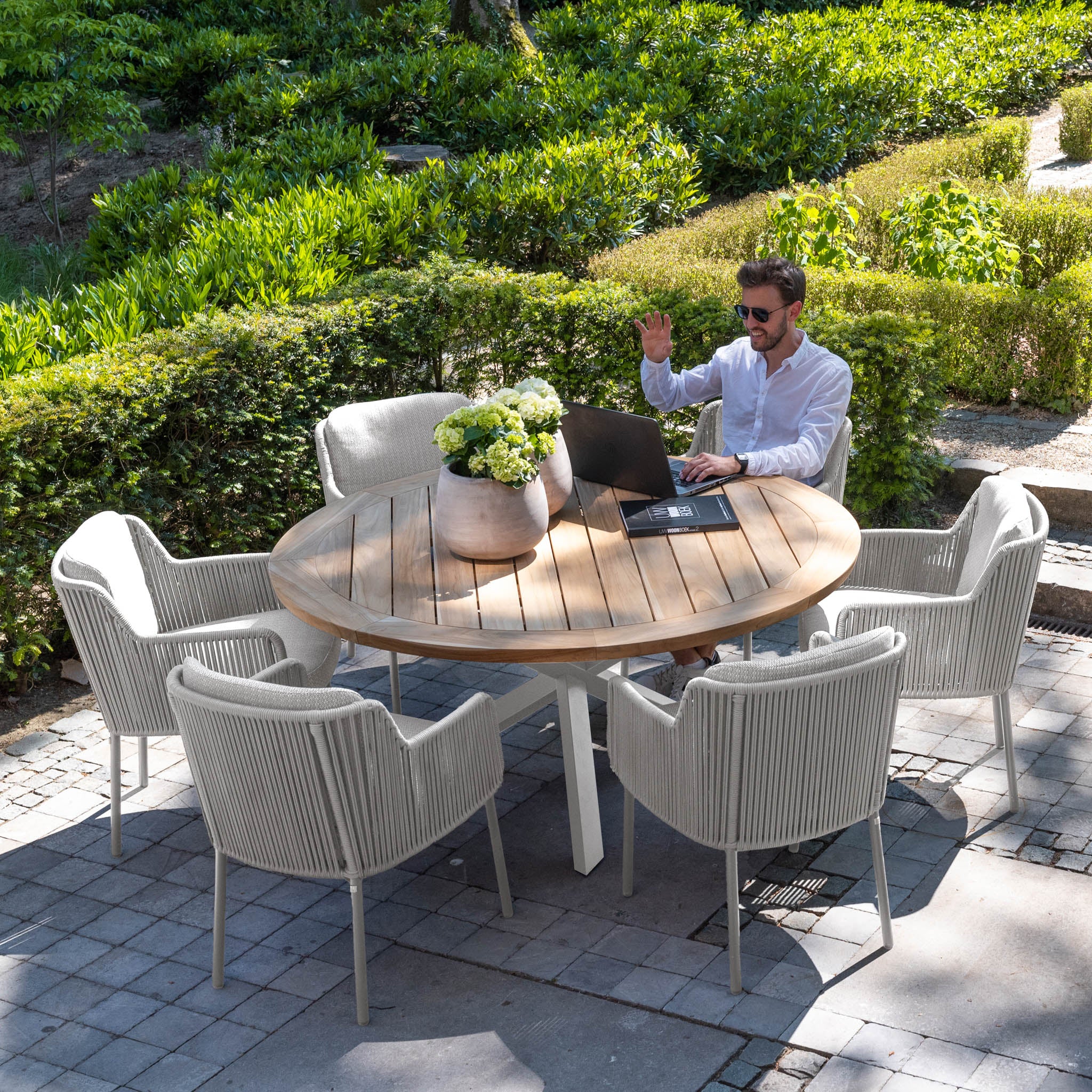 A person in sunglasses waves while sitting at a round outdoor table from the Bernini 6 Seat Round Dining with Teak Table in Light Grey range, with a laptop, surrounded by greenery.