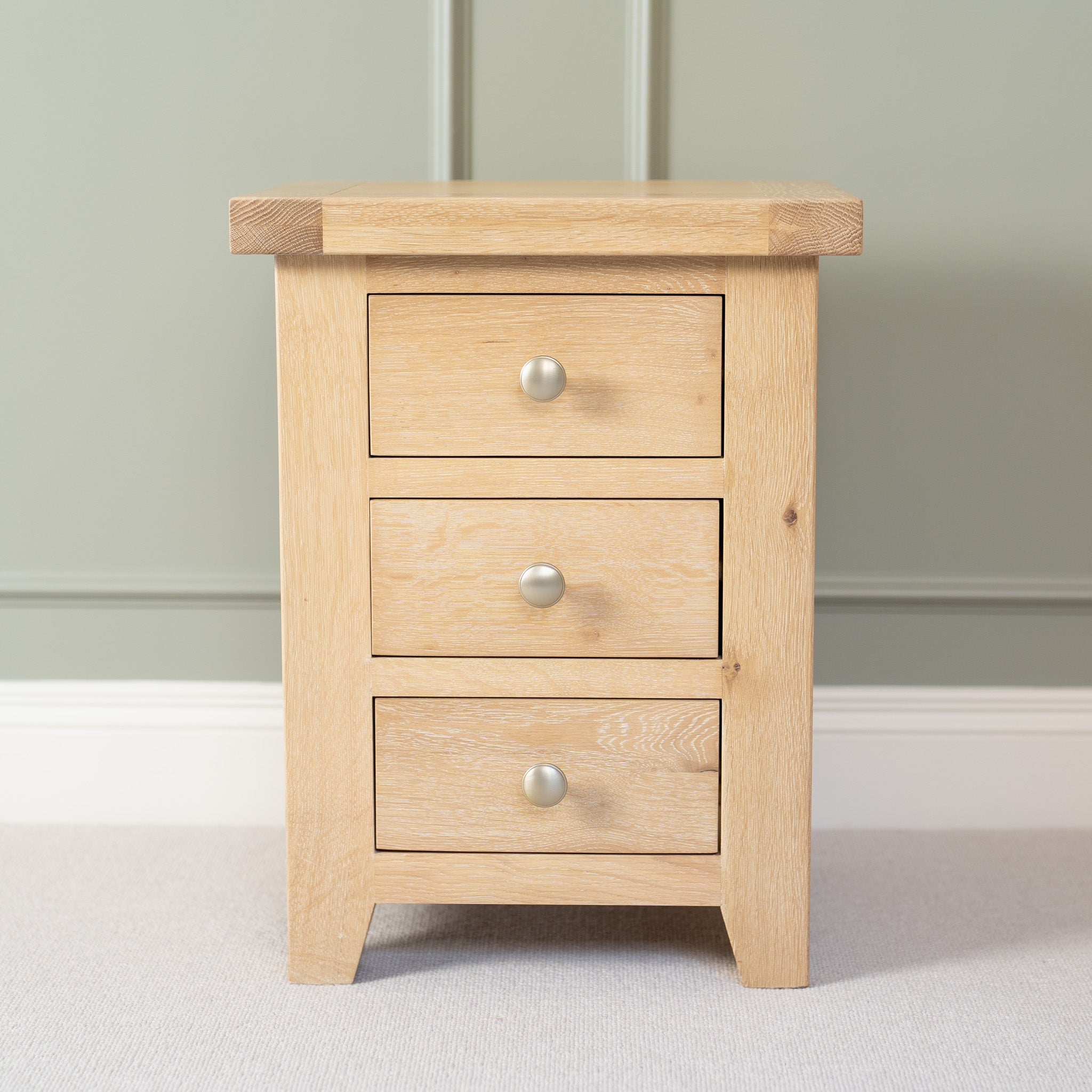 The Burford 3 Drawers Bedside Table in Natural Oak, adorned with silver knobs, stands elegantly against a green wall.