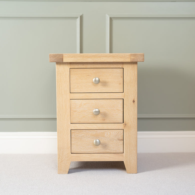 The Burford 3 Drawers Bedside Table in Natural Oak sits against a light green paneled wall on a carpeted floor, offering charm and versatility.