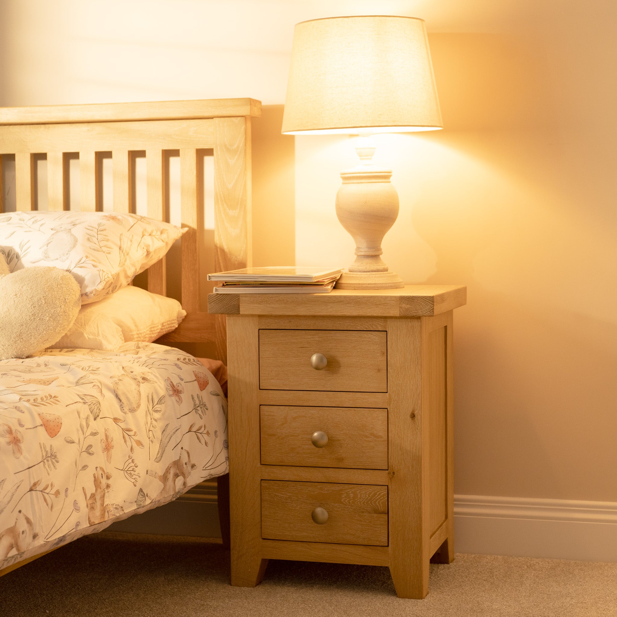 The Burford 3 Drawers Bedside Table in Natural Oak offers ample storage and complements floral bedding, complete with a lamp and books.