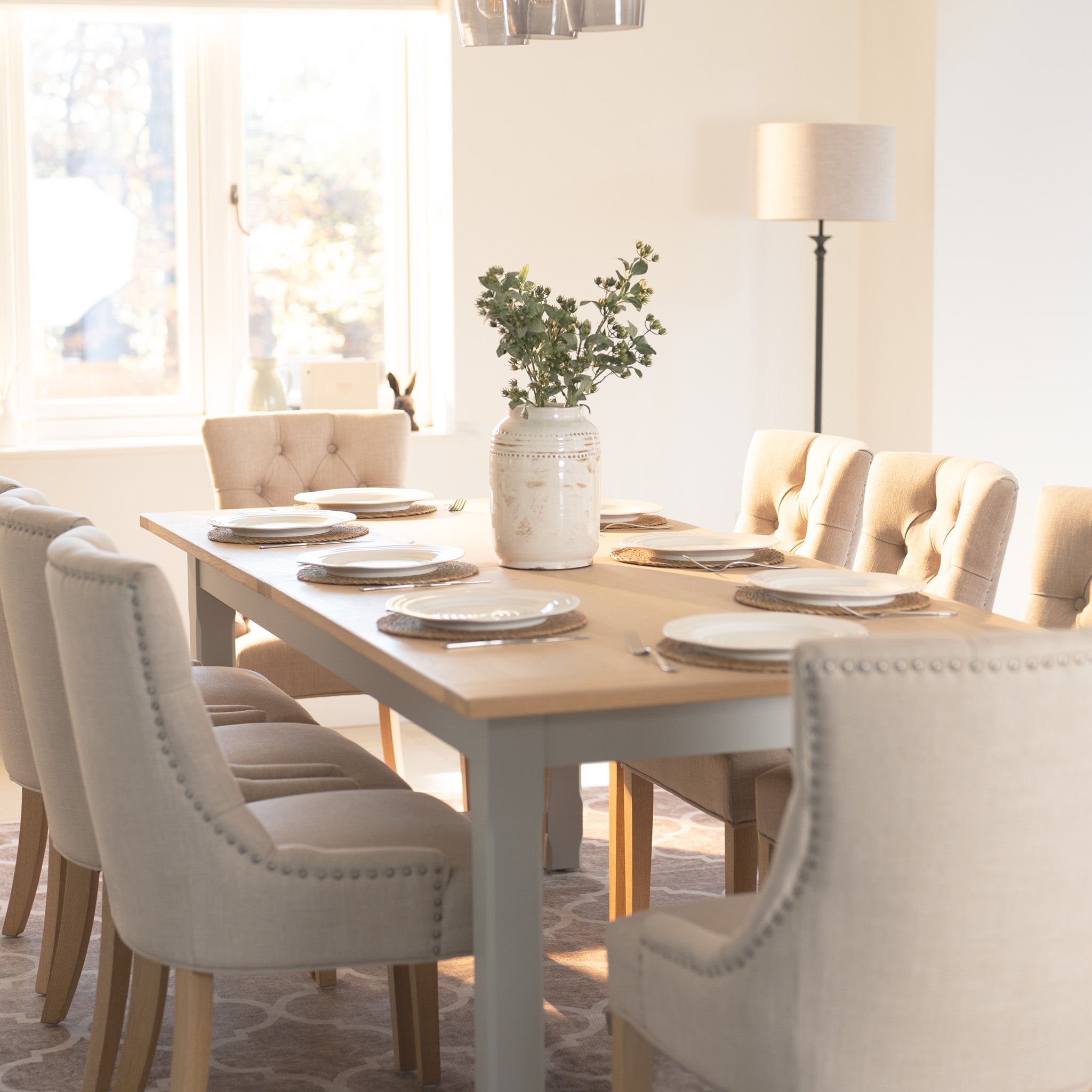 A bright dining room showcases the Burford 4-6 Seater Extending Dining Table in Pebble Grey, surrounded by beige chairs and set plates, complemented by a vase with greenery near a sunny window.