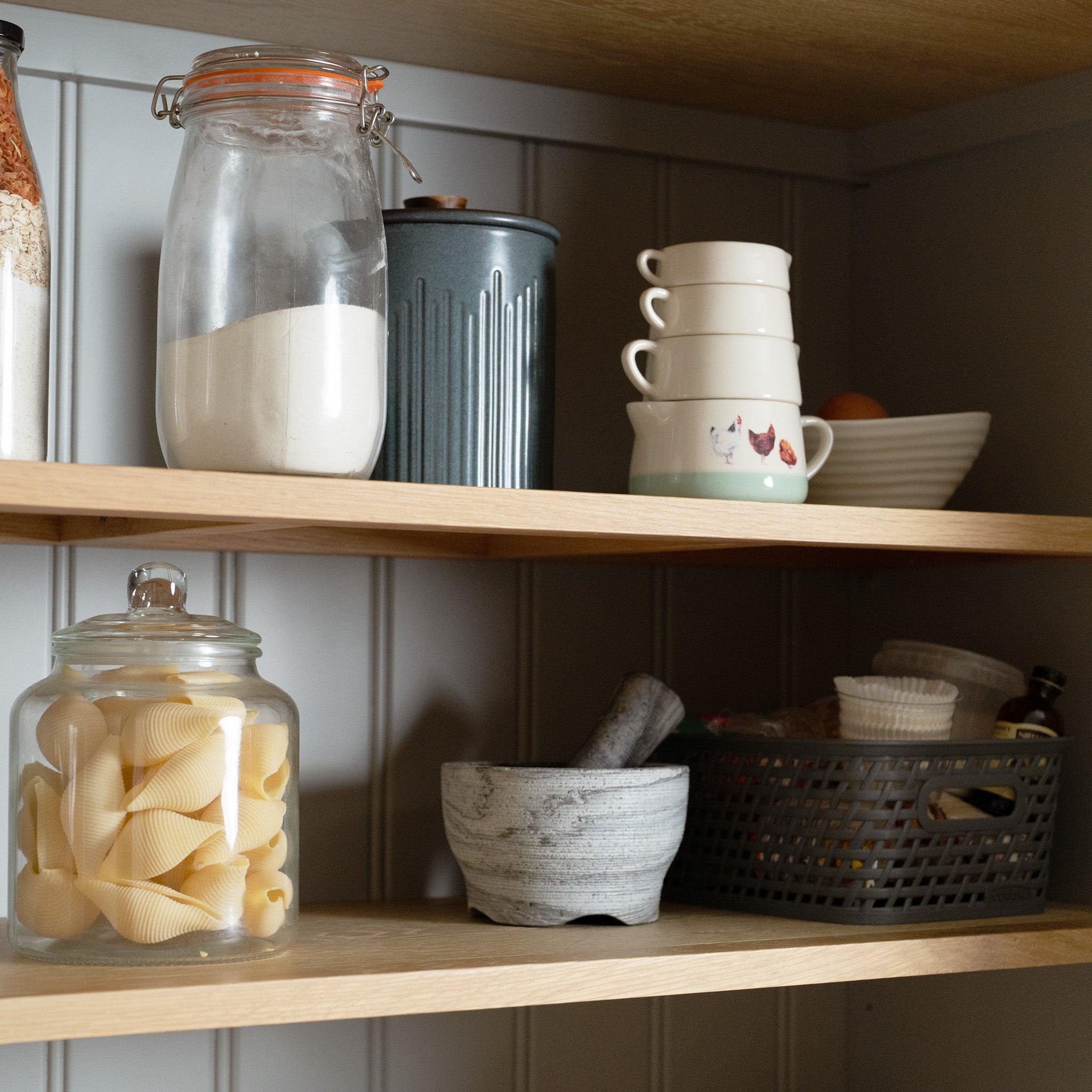 Pebble Grey wooden pantry shelves, like a Chalbury Triple Wardrobe, hold jars of pasta and flour, stacked cups, a mortar and pestle, and a basket with bottles, epitomizing organized charm.