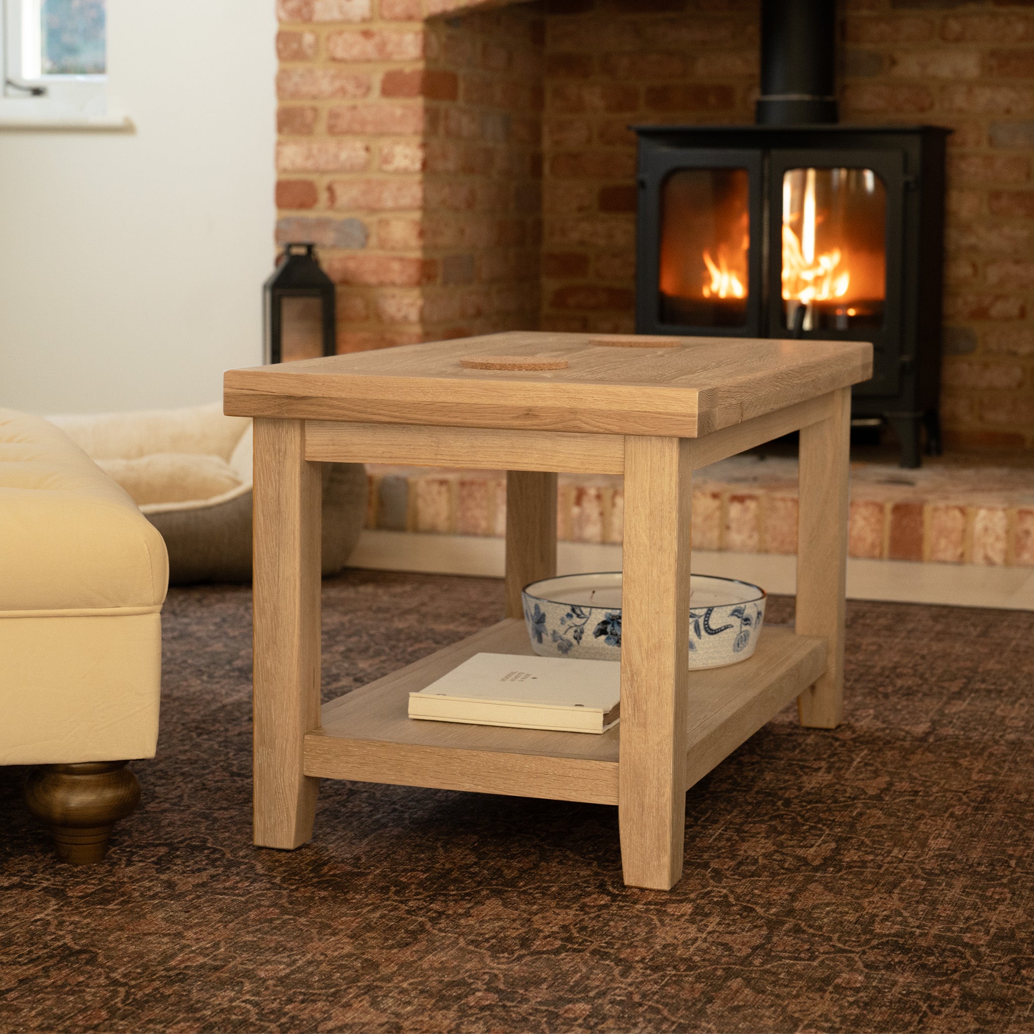 The Burford Large Coffee Table with Shelf in Natural Oak, tastefully adorned with books and a bowl, sits perfectly in front of a fireplace on a patterned rug.