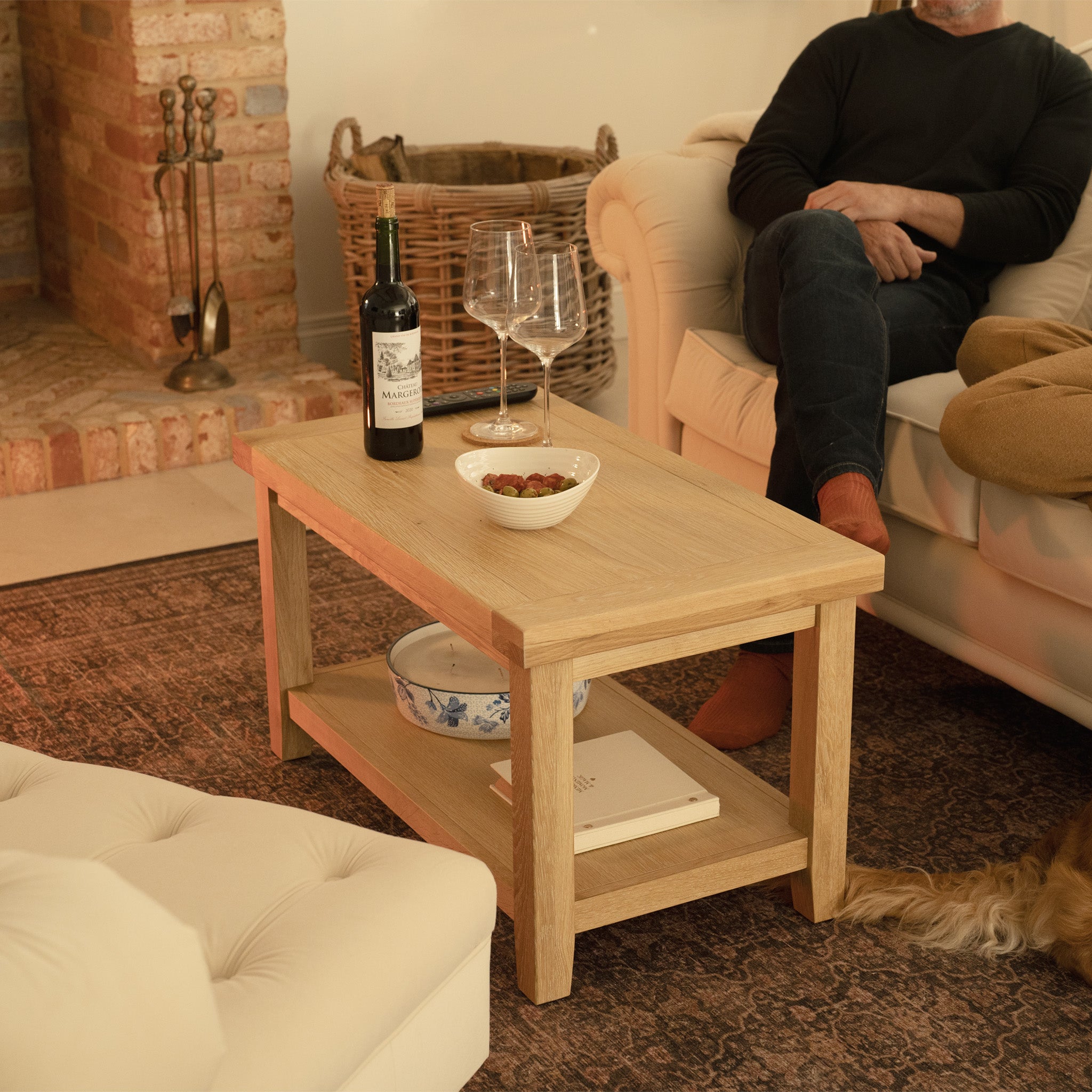 A cozy living room showcases a Burford Large Coffee Table with Shelf in Natural Oak. Wine, glasses, and snacks are invitingly placed near the fireplace where someone sits enjoying the warmth.