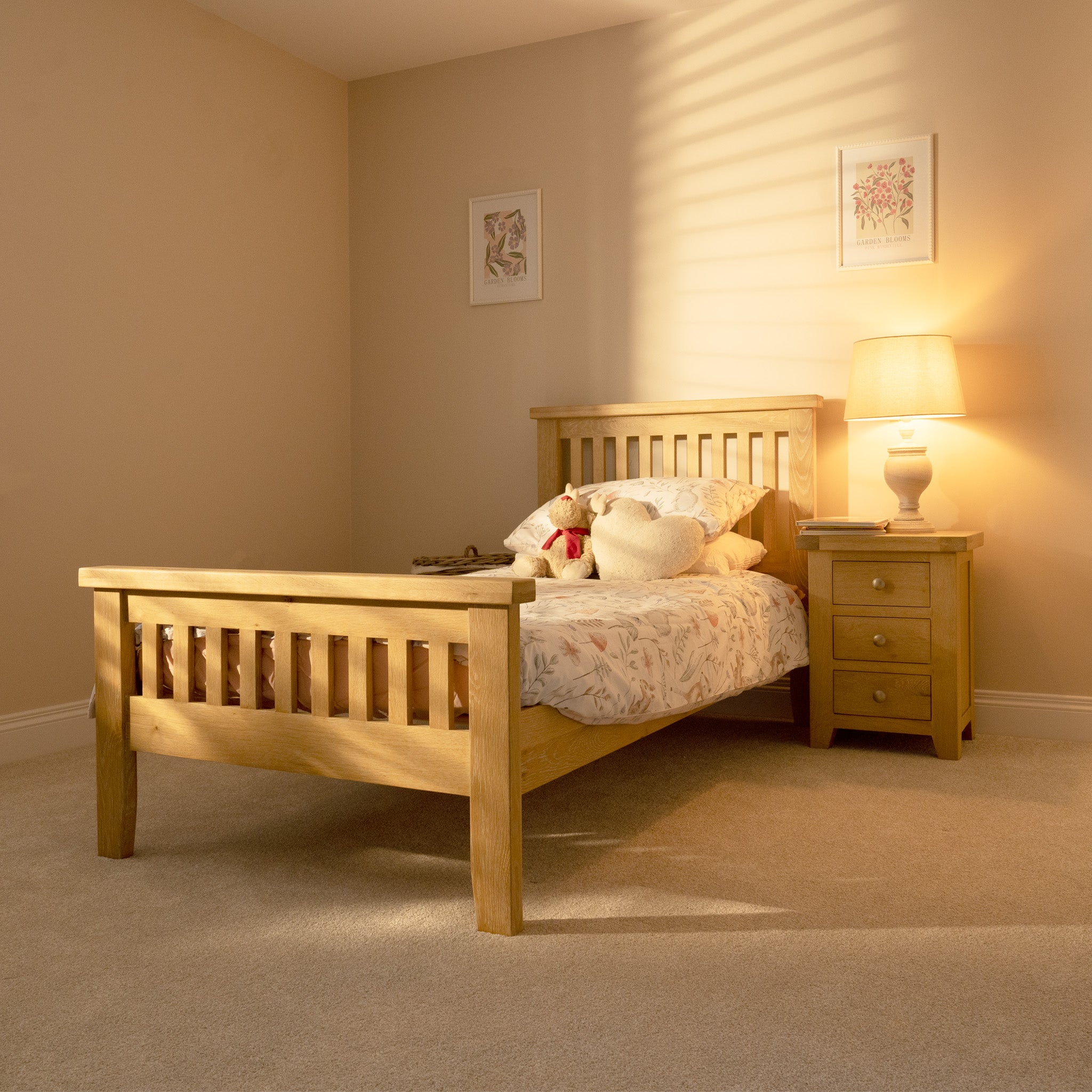 A cozy bedroom showcases the Burford 3ft High-End Single Bed Frame in Natural Oak, elegantly paired with heart-shaped pillows, a bedside table, lamp, and floral wall art. This tasteful blend of design elements and soft lighting crafts a warm oasis.