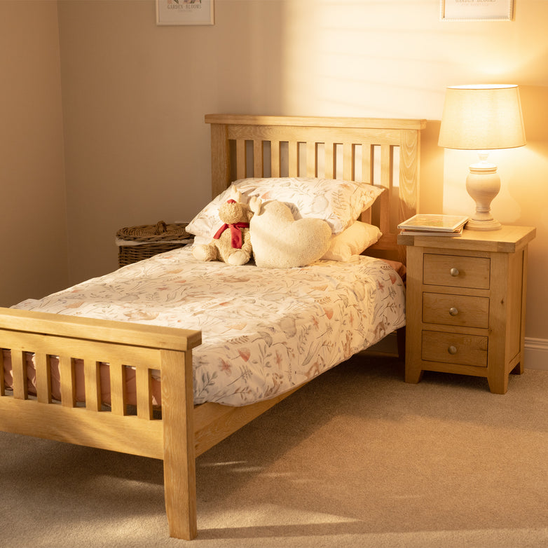 A cozy single bed with a Burford 3ft High-End Single Bed Frame in Natural Oak and floral bedding is complemented by a teddy bear and heart pillow. A wooden nightstand and lamp enhance the warmly lit room's design with natural oak accents.