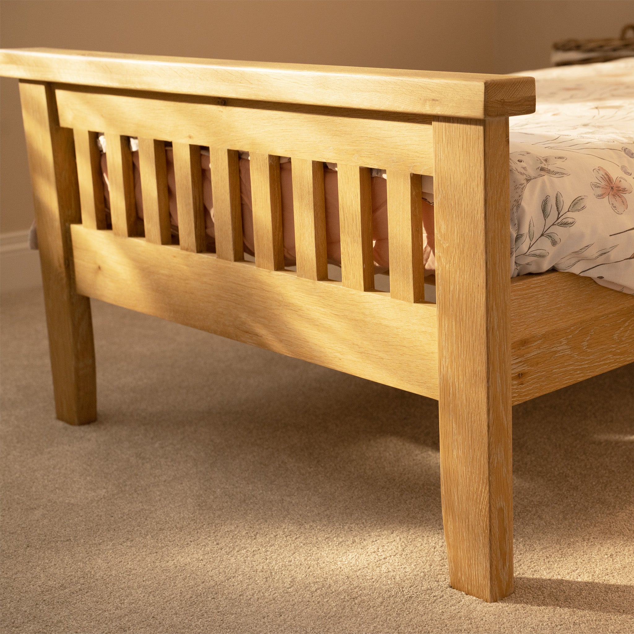 The Burford 3ft High-End Single Bed Frame in Natural Oak features a slatted headboard and floral bedspread, beautifully bathed in warm sunlight on a carpeted floor.