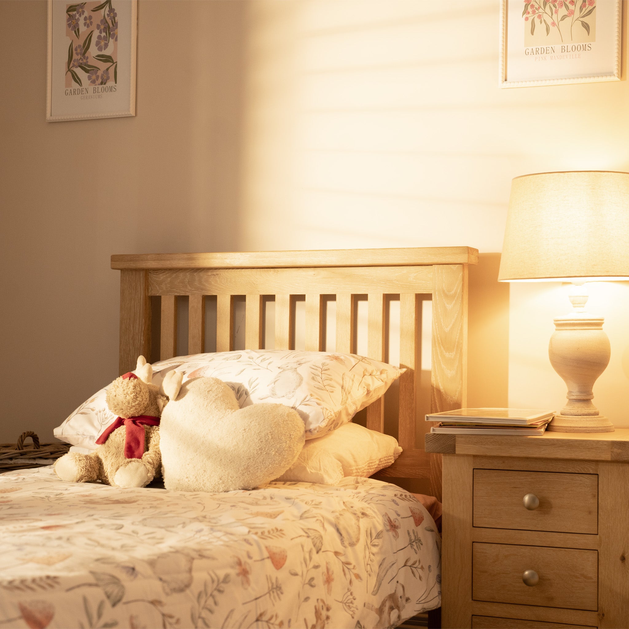 A cozy bedroom showcases a Burford 3ft High-End Single Bed Frame in Natural Oak, adorned with a heart-shaped pillow and teddy bear, while a lit lamp casts warm, inviting light on the nightstand.