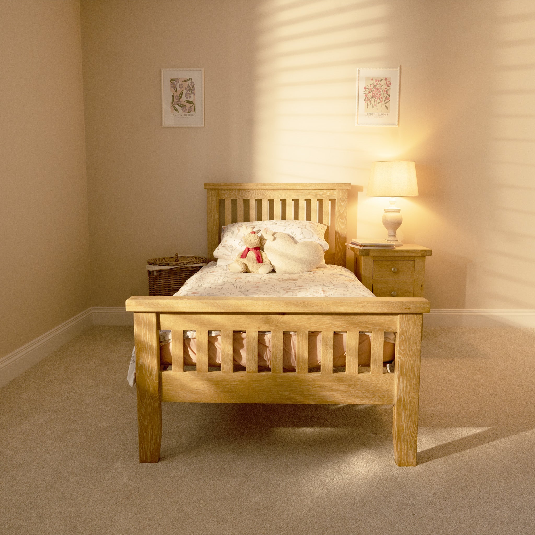 A cozy bedroom with a Burford 3ft High-End Single Bed Frame in Natural Oak, accompanied by a teddy bear, a nightstand with a lit lamp, and framed art on beige walls.