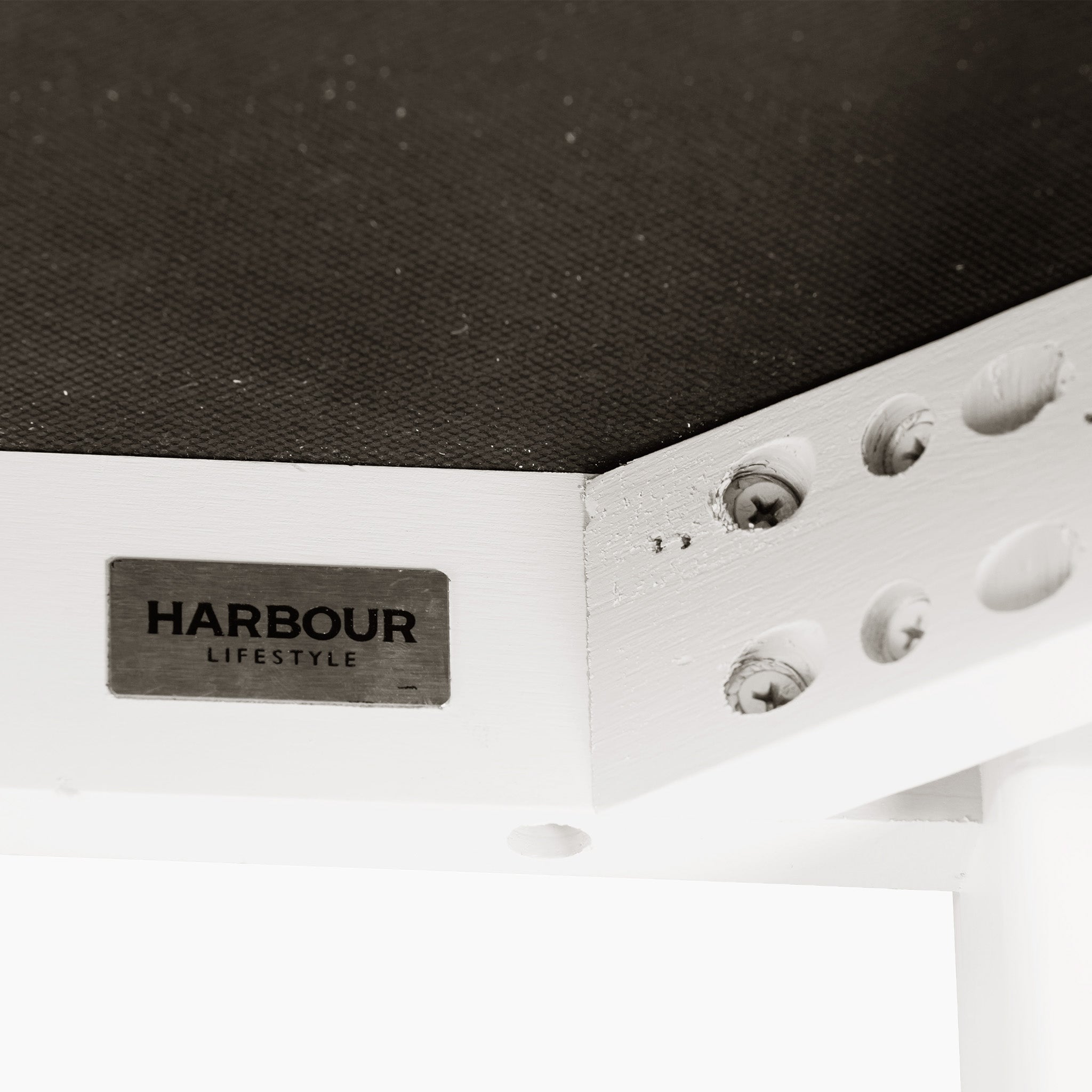 Close-up of a corner of a white table featuring a Harbor Lifestyle label, with visible metal bolts and an elegant design similar to the Chalbury Stool in Warm White.