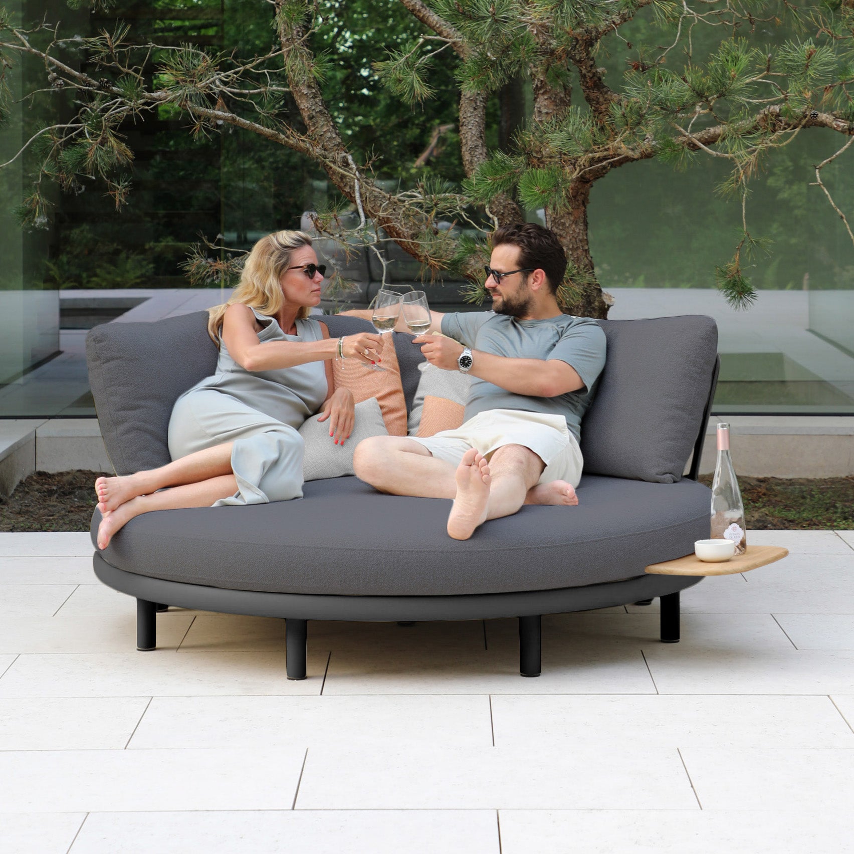 Two people relax on the weather-resistant Selene Daybed in Charcoal, drinks in hand, surrounded by lush greenery, epitomizing outdoor luxury.