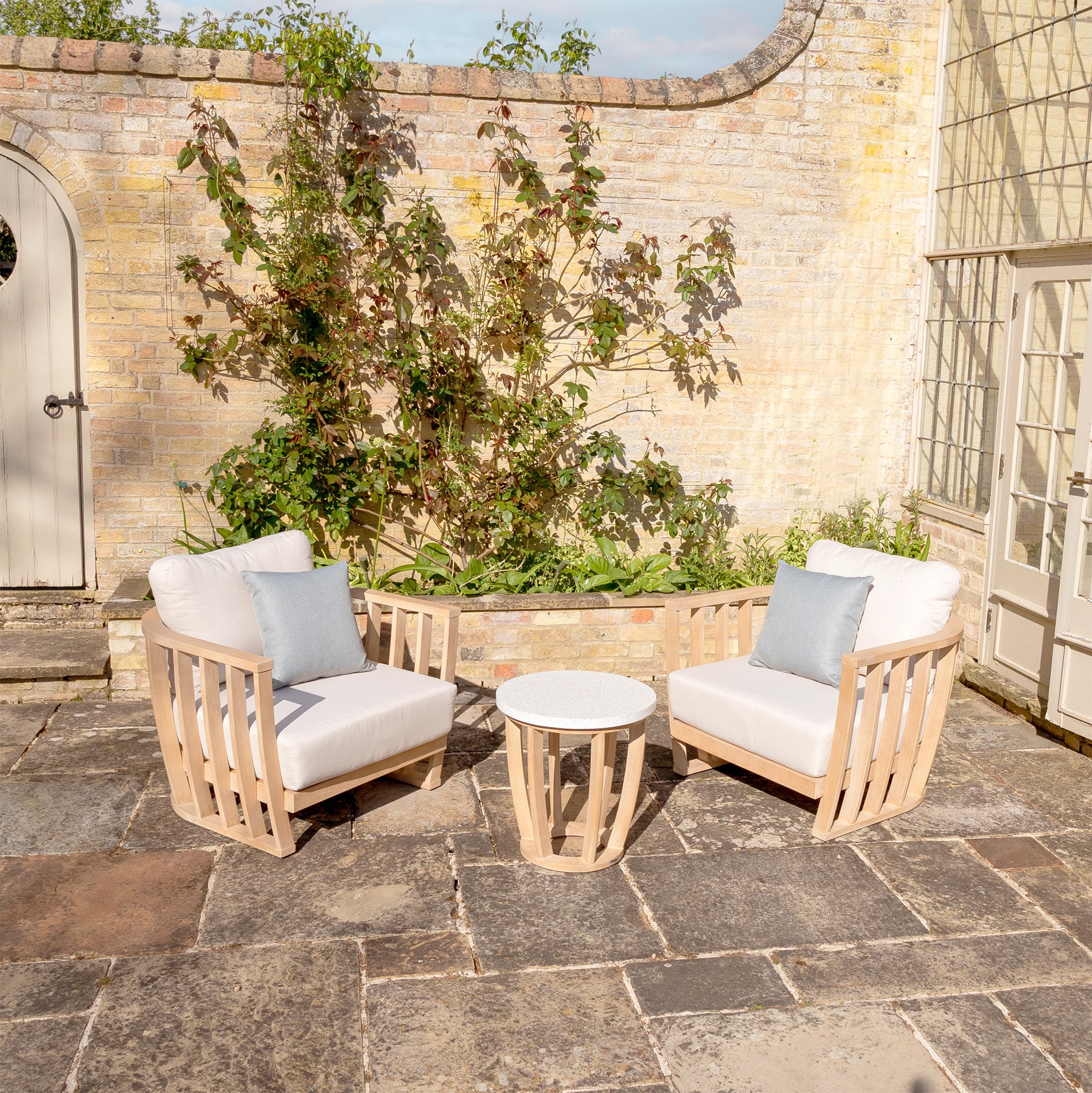 The Cape Lounge Set in Beige features two cushioned chairs with deep plush cushions and a small round table, all set against a brick wall adorned with greenery in the background. This inviting outdoor furniture set will transform any patio into a serene retreat.