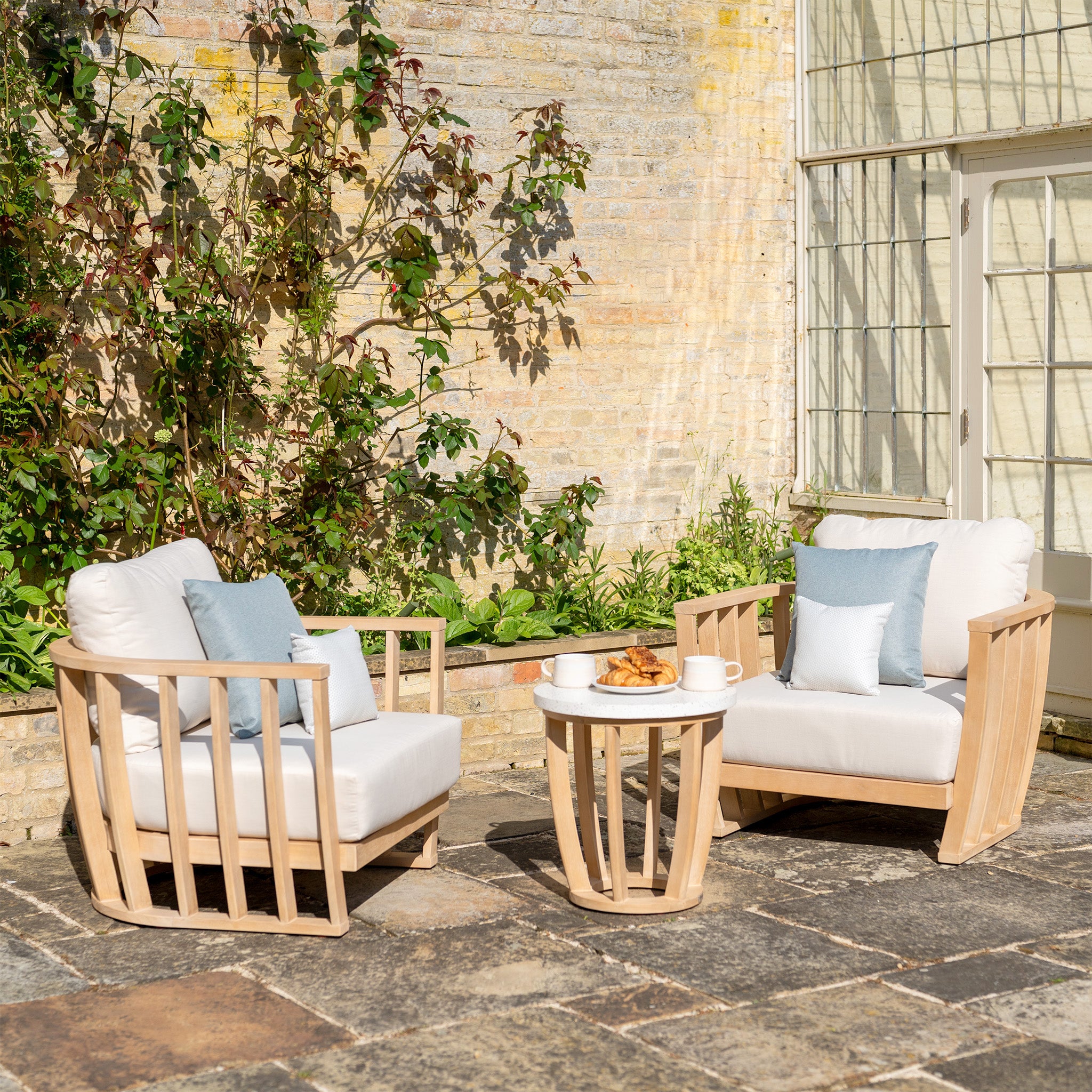 Outdoor seating area featuring the Cape Lounge Set in Beige, including two cushioned wooden armchairs and a small round table holding coffee cups and croissants, all set against a leafy backdrop.