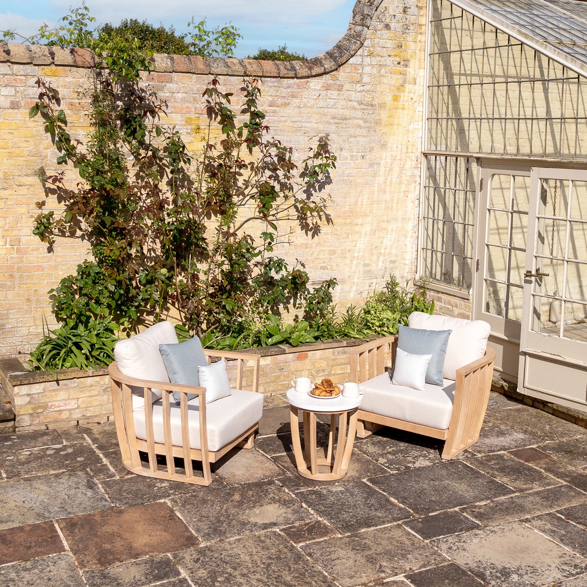 Outdoor patio showcasing a Cape Lounge Set in Beige, featuring two wooden armchairs with deep plush cushions, and a small round table holding a teapot and pastries, all situated near a brick wall.