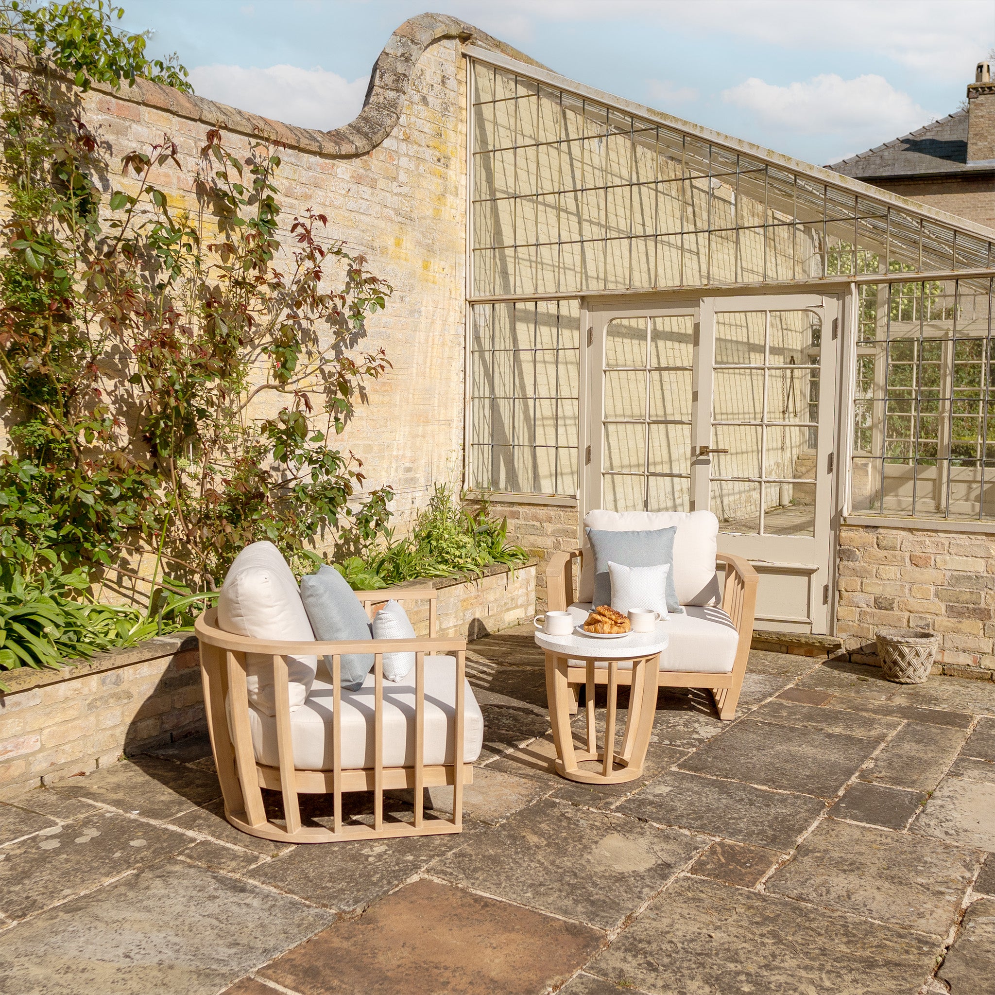 A patio featuring the Cape Lounge Set in Beige, which includes two deep plush cushioned chairs and a small table, set against a stone wall and a large glass greenhouse.