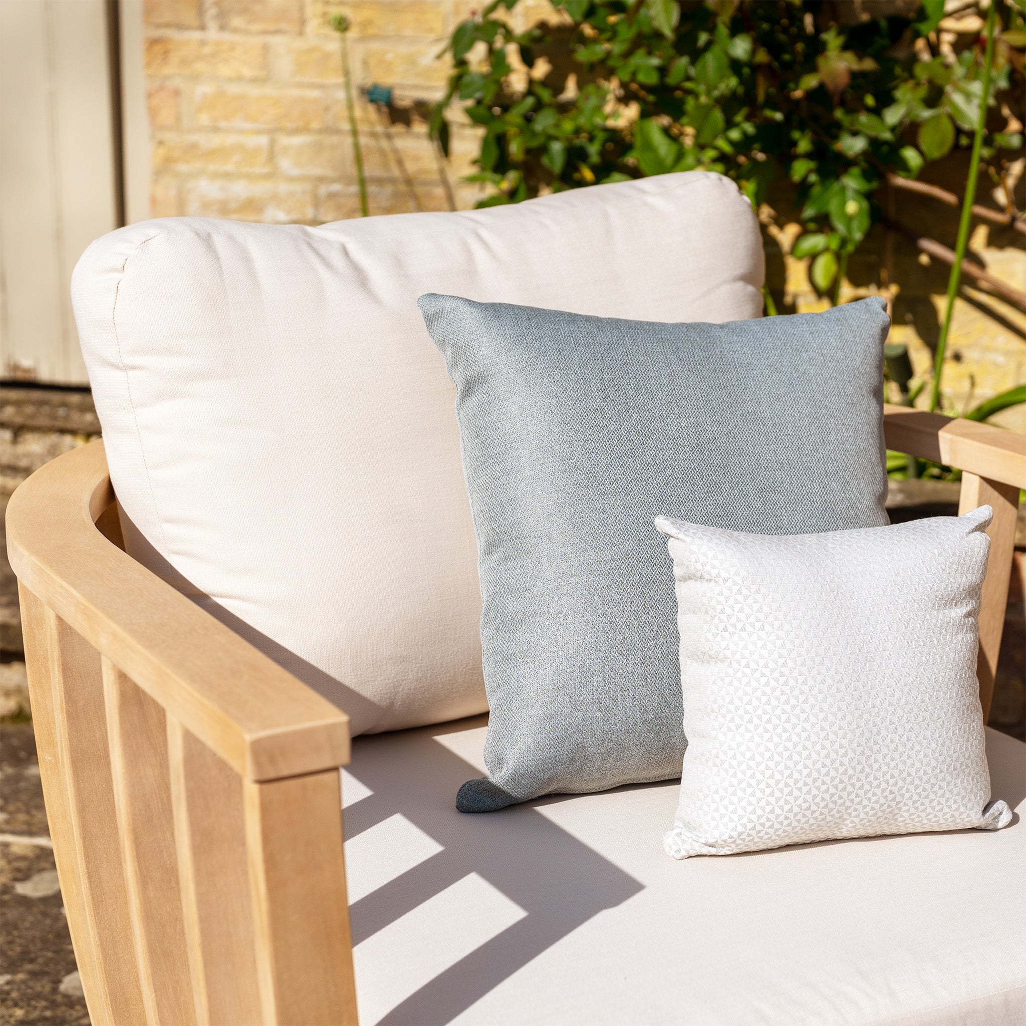 A wooden outdoor chair featuring deep plush white cushions, accompanied by grey and white pillows from the Cape Lounge Set in Beige, set against a brick wall backdrop.