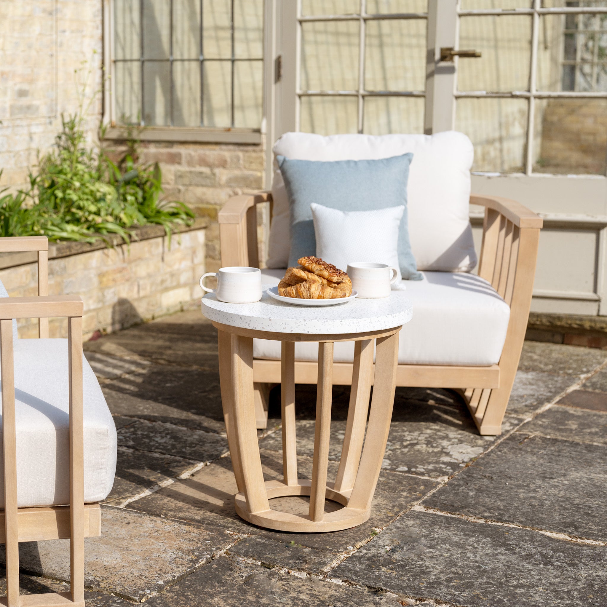 Outdoor seating area features the Cape Lounge Set in Beige with wooden furniture, deep plush cushions, and a small table holding two mugs and a plate with pastries.