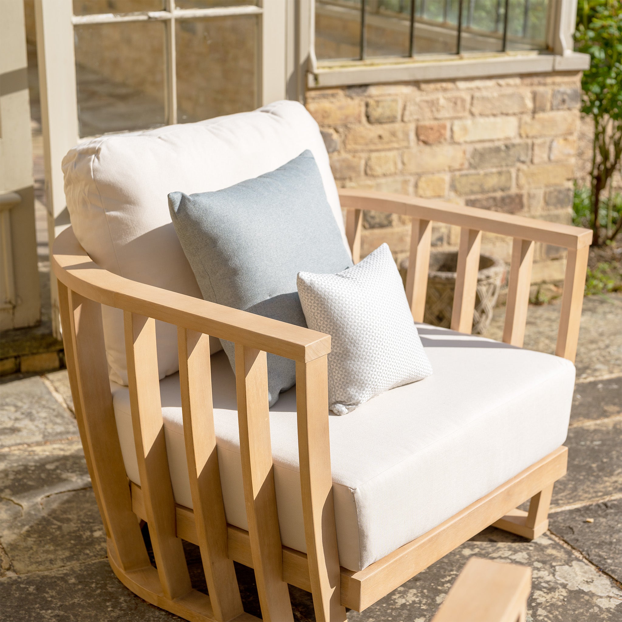 A Cape Lounge Set in Beige, featuring a wooden armchair with deep plush cushions and two throw pillows, is placed on a stone patio in front of a brick wall with windows.