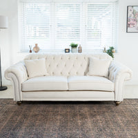 In a bright room with decorative items, the Chesterfield 3 Seat Velvet Sofa in Cream sits elegantly on a patterned rug, adorned with plush cushions and bordered by blinds.