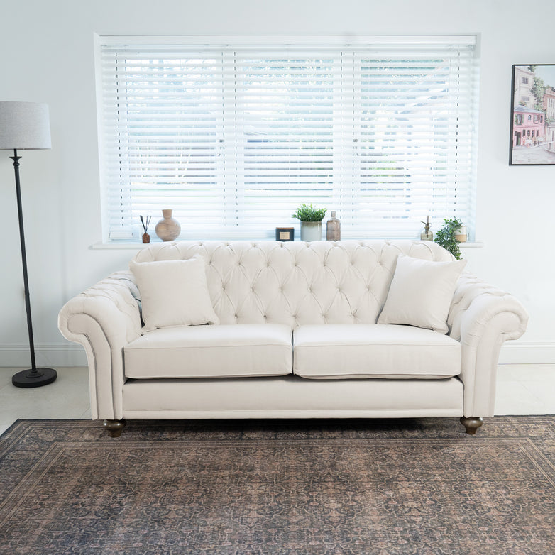 The Chesterfield 3 Seat Sofa in Ivory, boasting a traditional British tufted design, sits with cushions in a bright room featuring a lamp and window blinds.