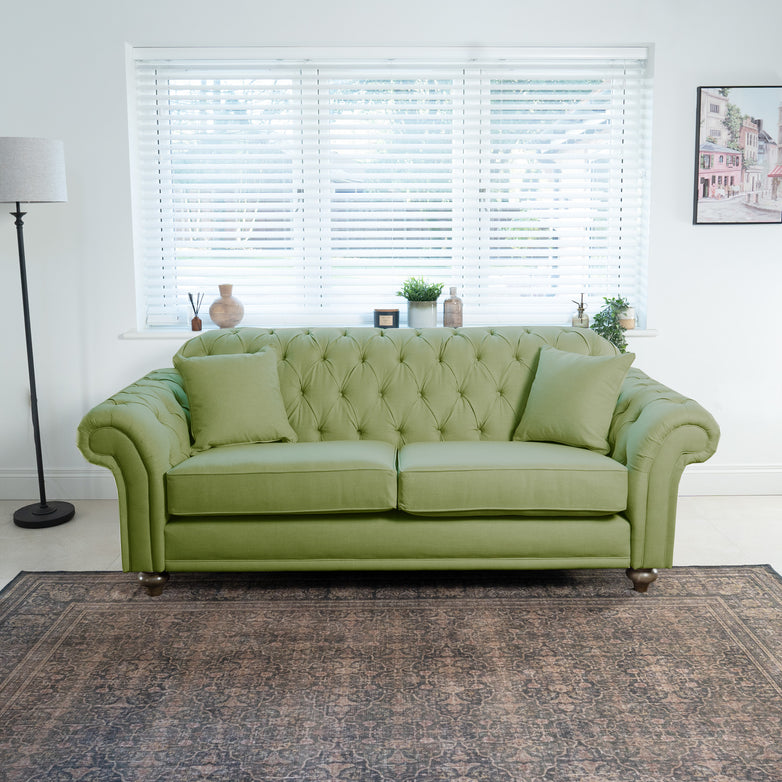 A Chesterfield 3 Seat Sofa in Olive with cushions is situated in a bright living room, complete with blinds, a rug, lamp, and wall art. The olive color highlights British craftsmanship, bringing timeless elegance to the space.