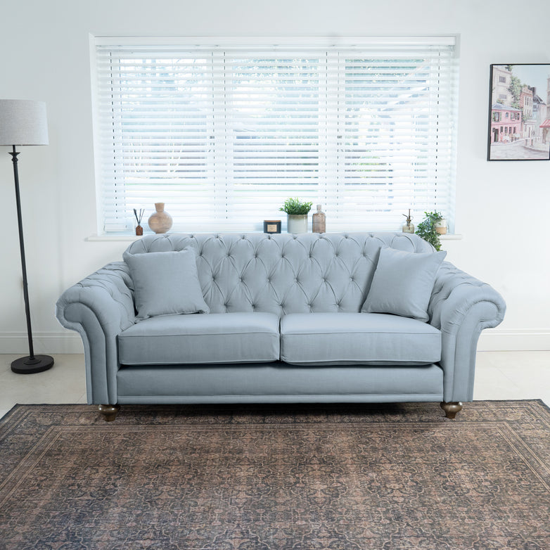 A Chesterfield 3 Seat Sofa in Sky Blue, complete with pillows, is situated in a bright room featuring a window, floor lamp, and a framed picture on the wall.