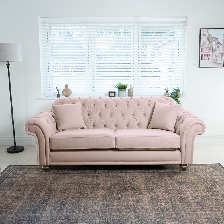 In a bright room, the dusky pink tufted Chesterfield 3 seat sofa showcases British craftsmanship, with cushions resting elegantly. Window blinds stand behind, while plants and a lamp gracefully flank either side.