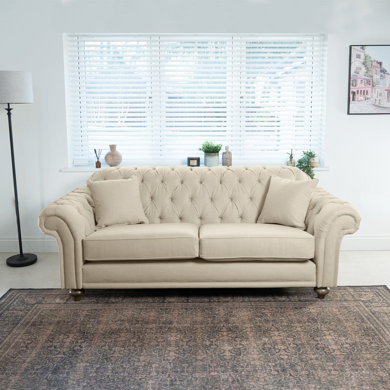 Handcrafted in England, the Chesterfield 3 Seat Sofa in Beige is elegantly placed in a bright room with cushions, a chic rug, a stylish lamp, and framed art on the wall.