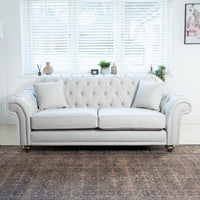 The warm grey, handcrafted Chesterfield 3 seat sofa with two cushions sits elegantly in a bright room featuring a large window and decorative rug, highlighting the beauty of sustainable materials.