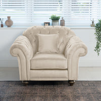 The Chesterfield Velvet Armchair in Cream, handcrafted in England from sustainable birch and beech, with a cushion, sits elegantly in front of a window with blinds and decor items on the windowsill.