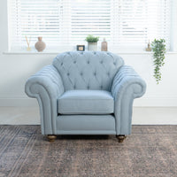 A Chesterfield Armchair in Sky Blue enhances a bright room with white blinds and decorative windowsill items. Its elegant tufted design is complemented by Dacron foam core for added comfort.