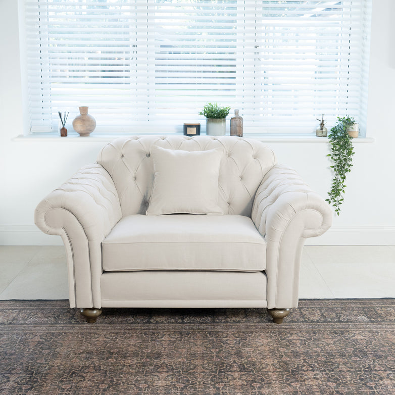 A beige tufted armchair with a cushion sits in front of a window with blinds and small potted plants, perfectly complementing the nearby ivory Chesterfield Loveseat.